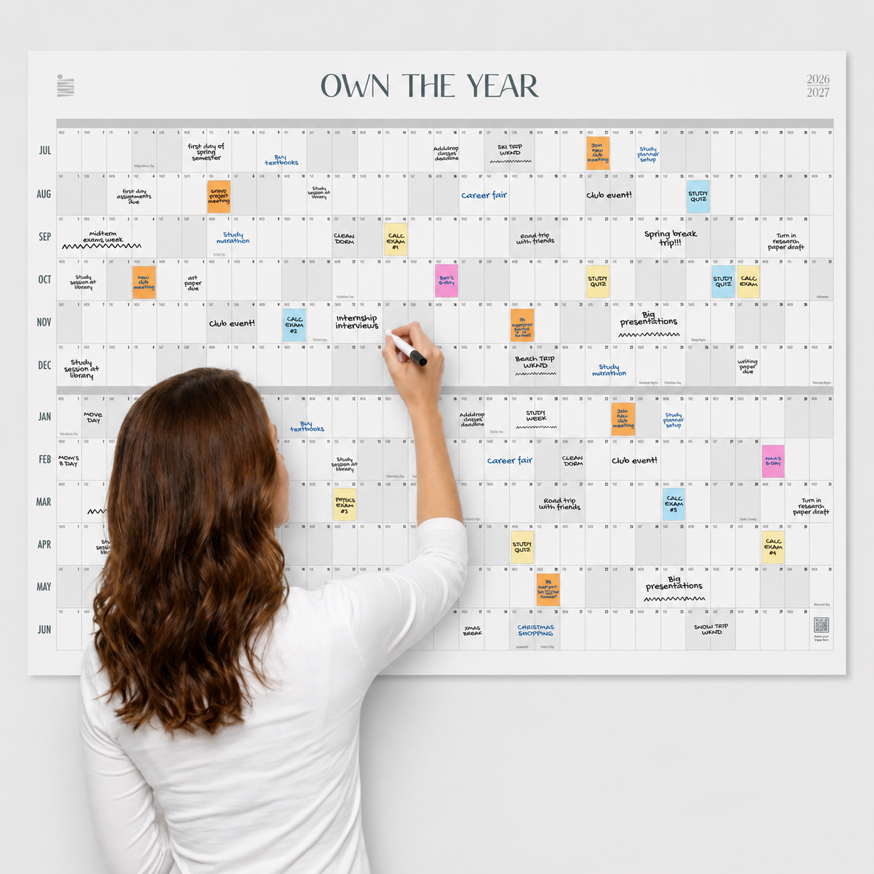 A woman writes on the Eterna Goods 2026-2027 Midyear/Academic Own The Year Calendar, a large wall planner covered in colorful notes and events from July to June.