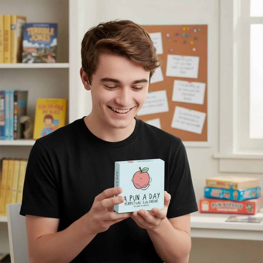 A young man in a black shirt smiles while holding the rockdoodles "A Pun A Day Perpetual Calendar"—a fun wordplay gift—in a room with shelves of books, games, and a corkboard in the background.