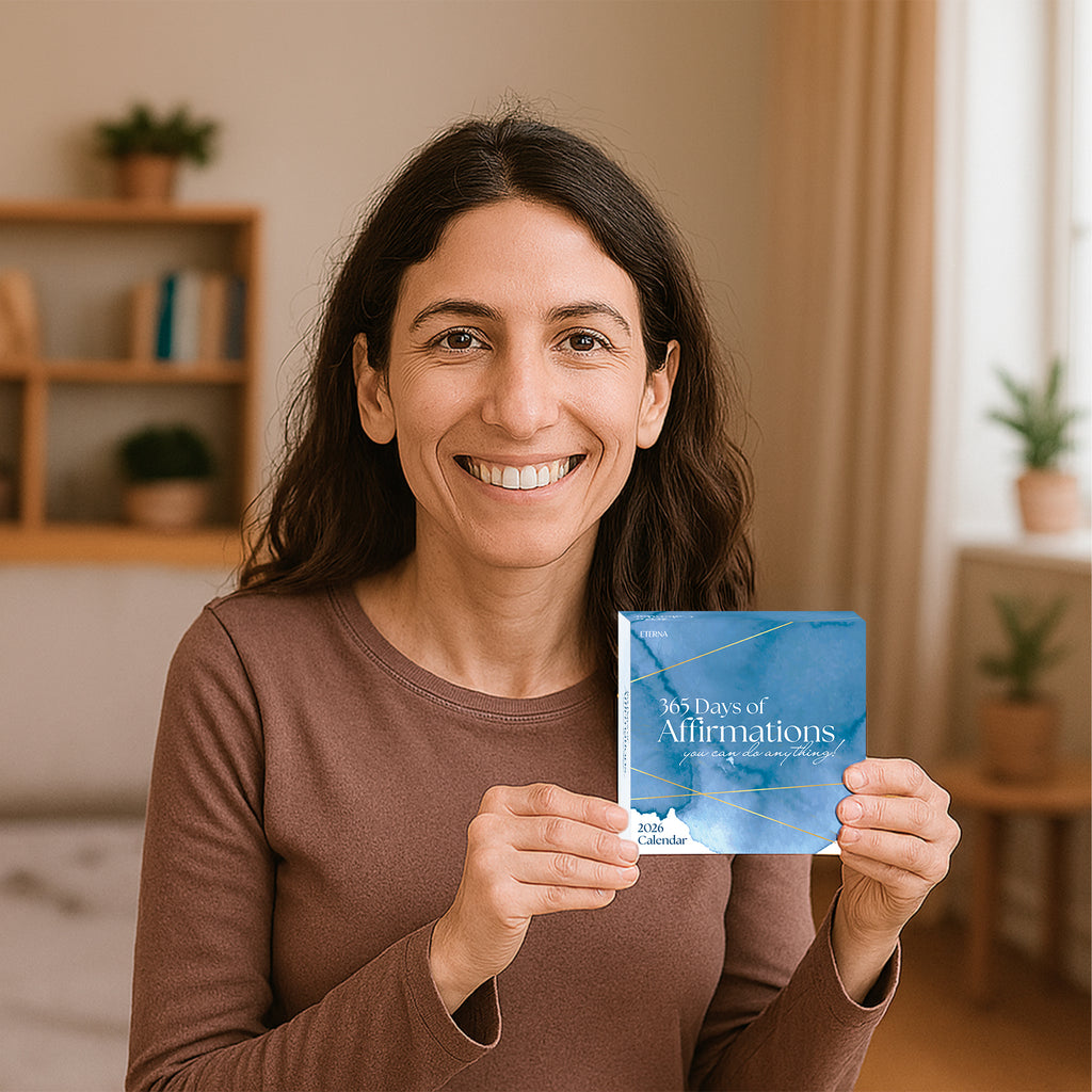 A woman smiles as she holds the Eterna Goods "2026 Daily Affirmations Calendar" in a softly lit room with plants and shelves, excited to enjoy her daily tear-off affirmation pages.