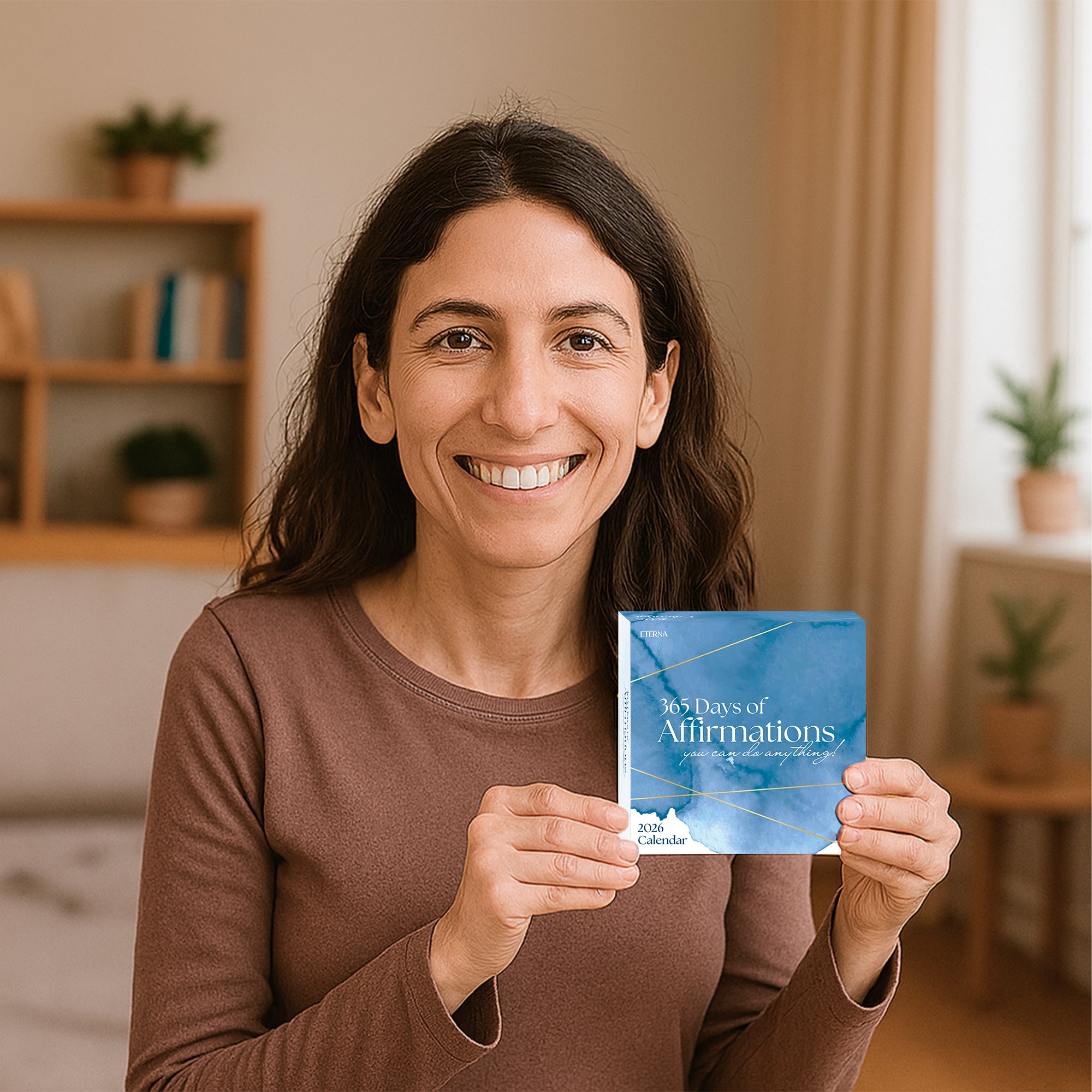 A woman smiles as she holds the Eterna Goods "2026 Daily Affirmations Calendar" in a softly lit room with plants and shelves, excited to enjoy her daily tear-off affirmation pages.