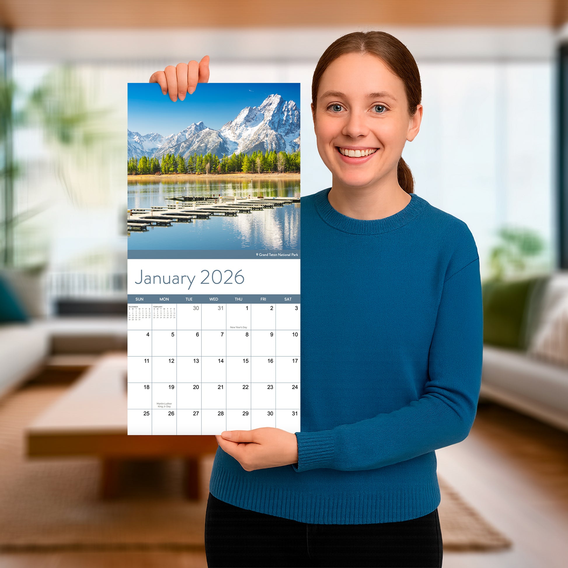 A woman in a blue sweater holds up the Eterna Goods 2026 National Parks Wall Calendar, featuring a scenic mountain and lake photo for January, in a modern living room.