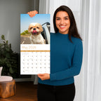 A woman in a blue sweater holds up the Eterna Goods 2026 Dogs Wall Calendar, opened to May and showing a dog with its tongue out, sticking its head out a car window.