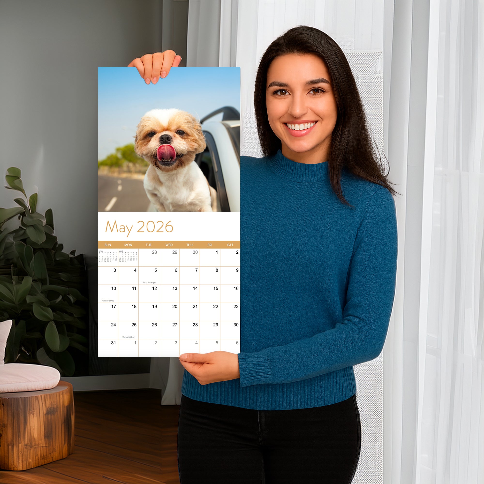 A woman in a blue sweater holds up the Eterna Goods 2026 Dogs Wall Calendar, opened to May and showing a dog with its tongue out, sticking its head out a car window.