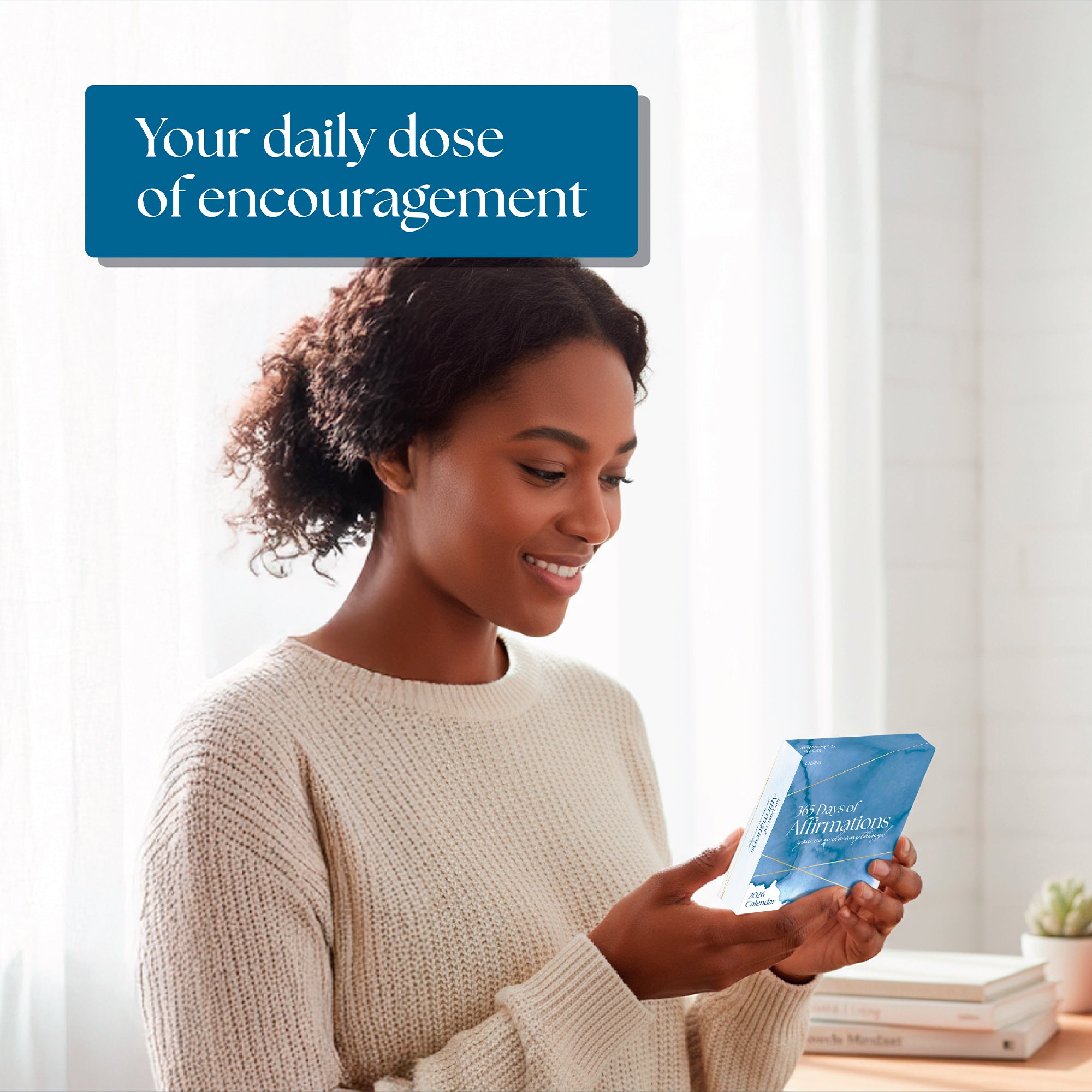 A woman in a cream sweater smiles while reading the blue “2026 Daily Affirmations Calendar” by Eterna Goods in a bright, softly lit room. Text above reads, “Your daily dose of encouragement.”.