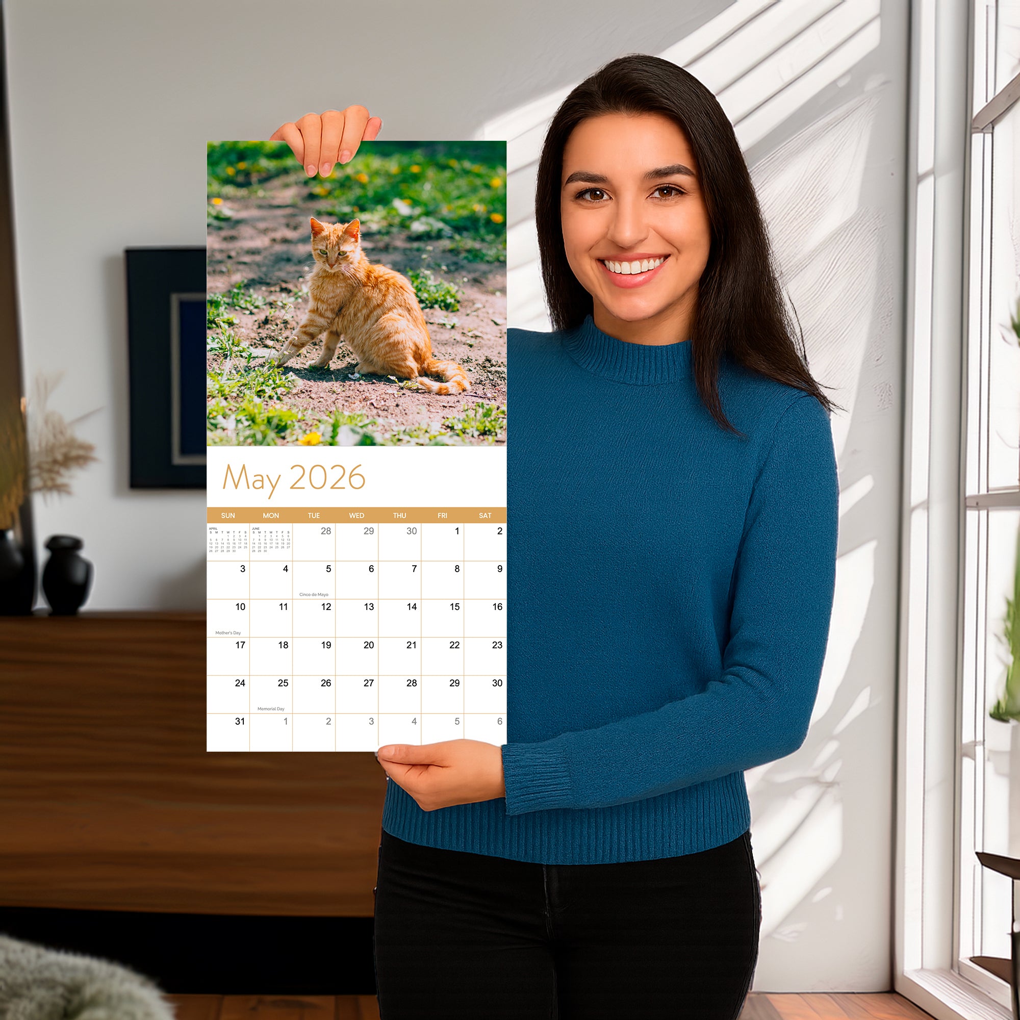A woman in a blue sweater stands indoors, smiling and holding an Eterna Goods 2026 Cats Wall Calendar with a cover photo of an orange cat outdoors.