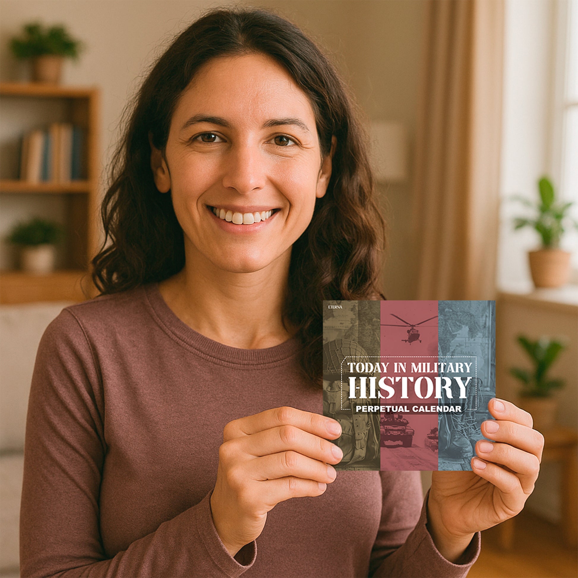 A woman smiles as she holds the "Today In Military History Perpetual Calendar" by Eterna Goods in a bright room with shelves and potted plants in the background.