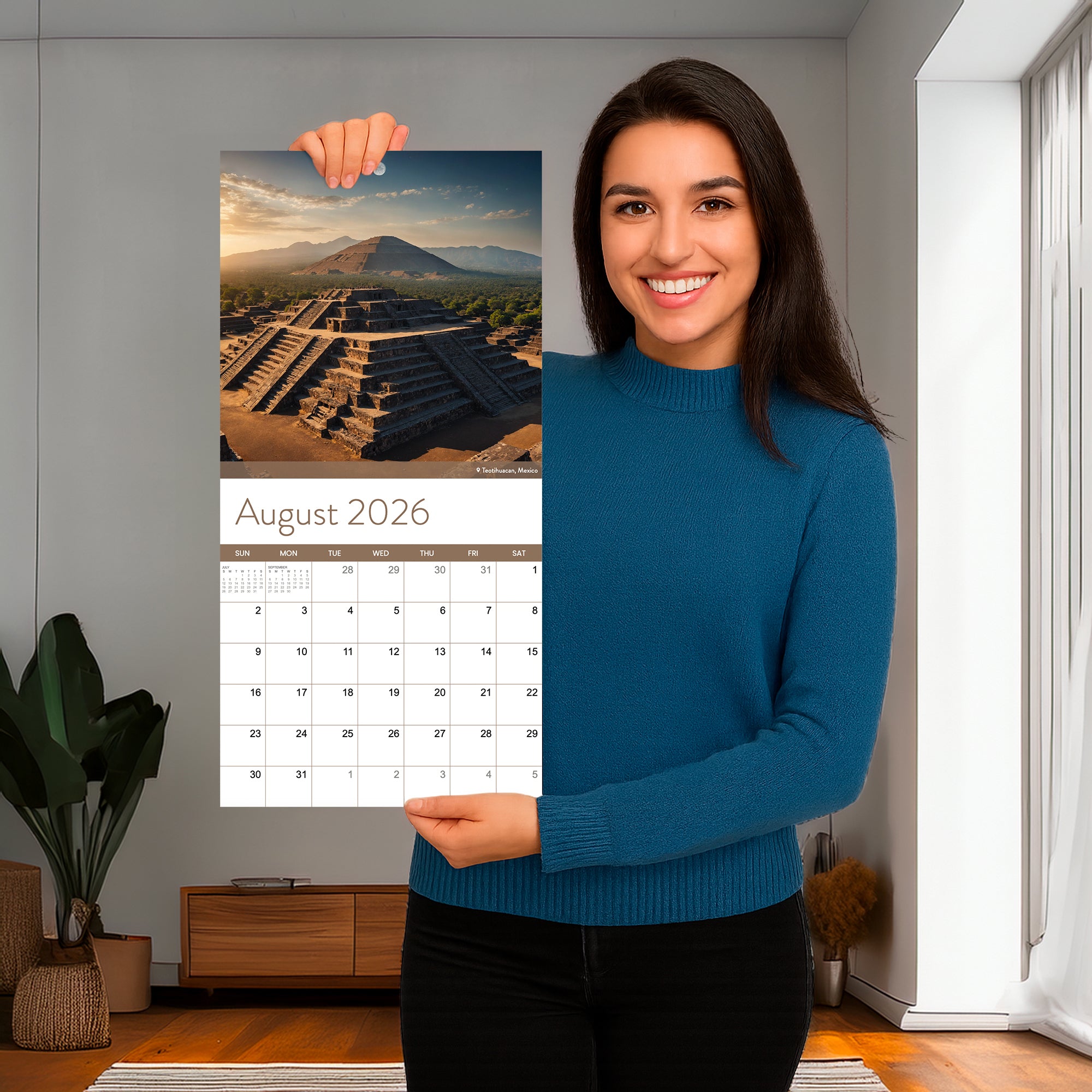 A woman in a blue sweater holds up the Eterna Goods 2026 Travel Inspiration Wall Calendar, opened to August, displaying a photo of an ancient stepped pyramid.