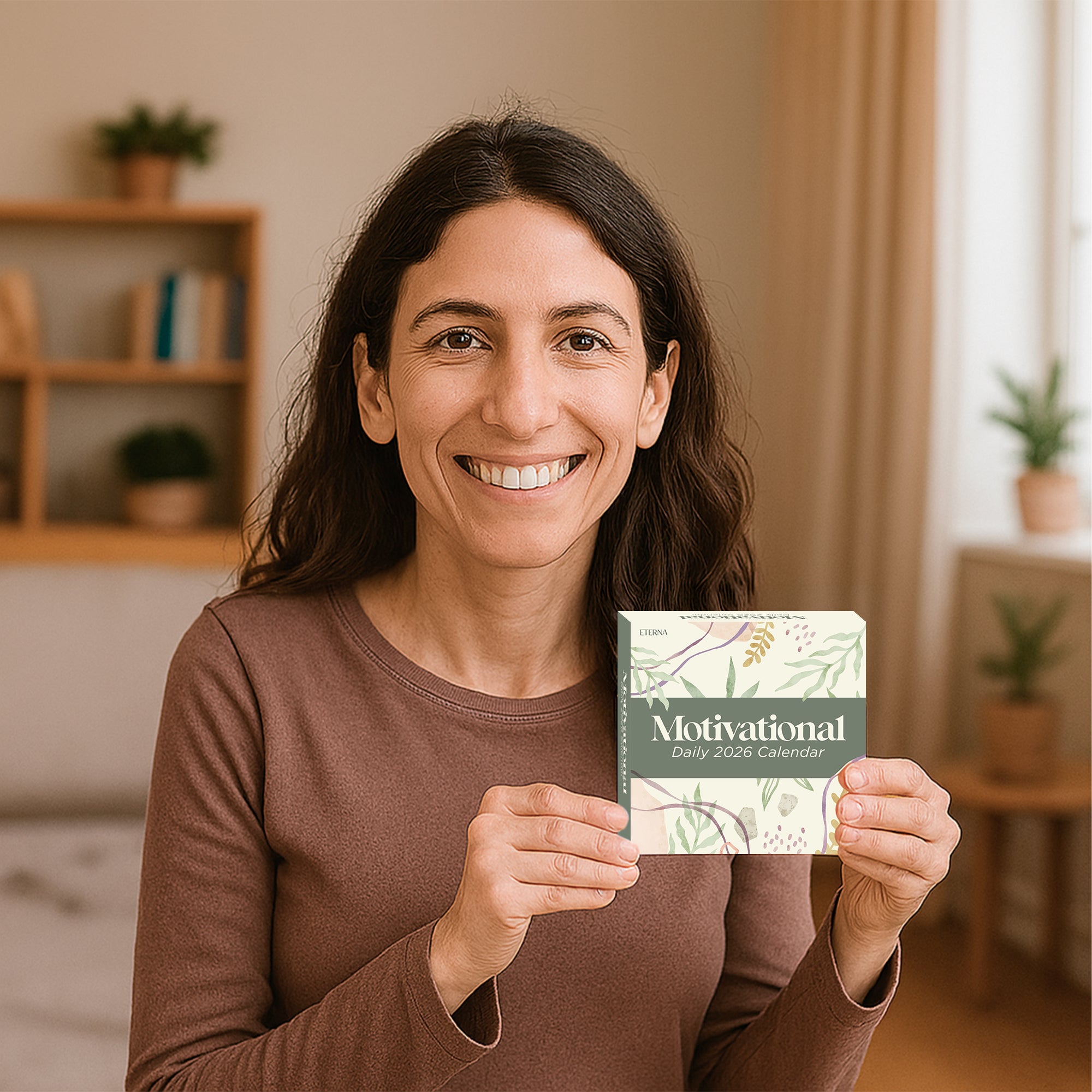 A woman smiles while holding the Eterna Goods 2026 Motivational Calendar, a tear-off desk calendar with daily motivational messages, in a cozy room decorated with plants and wooden shelves.