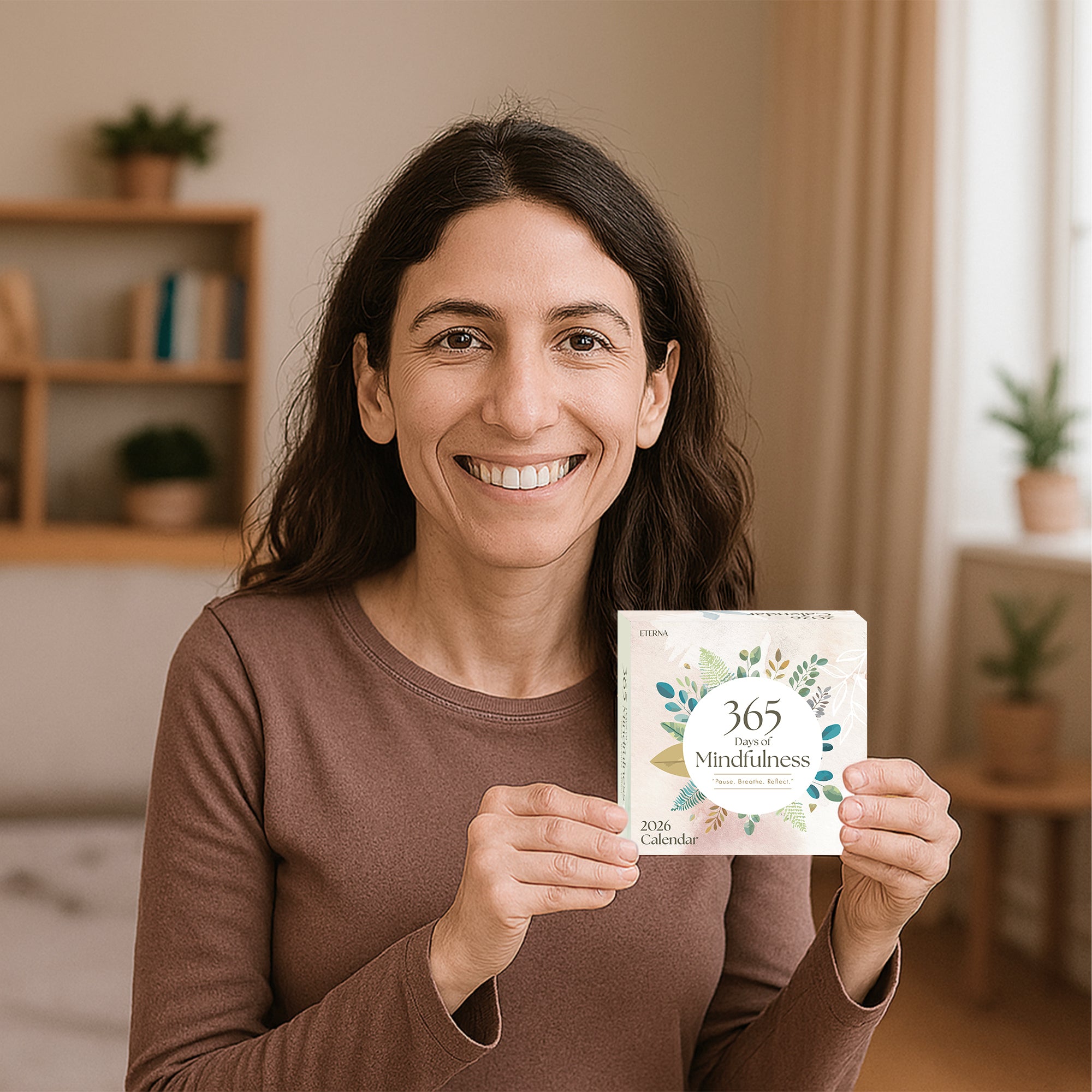 A woman smiles while holding the "2025 Mindfulness Calendar" by Eterna Goods in a cozy room with plants and shelves—a perfect daily mindfulness gift.