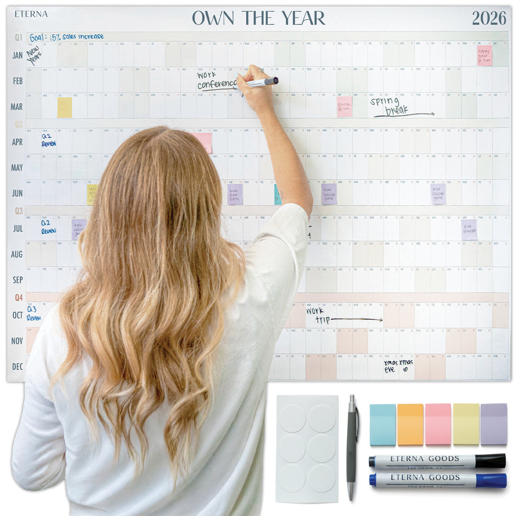 A woman writes on Eterna Goods' Own the Year 2026 GIANT Dry Erase Wall Calendar (4ft x 3ft) with markers, pen, eraser, and magnets—an ideal yearly planner for staying organized all year.
