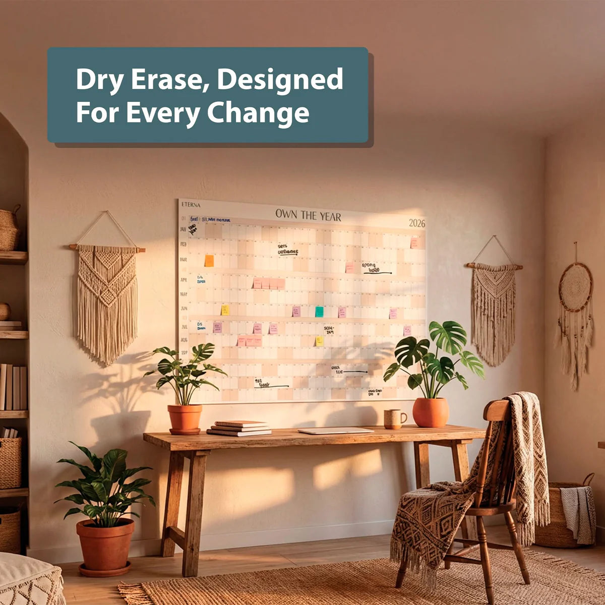 A home office with plants, a wooden desk, chair, and the Own The Year Giant Dry Erase Calendar by Eterna Goods serving as a large wall calendar and dry erase surface; the wall reads, "Dry Erase, Designed For Every Change." Macrame decor hangs on both sides.