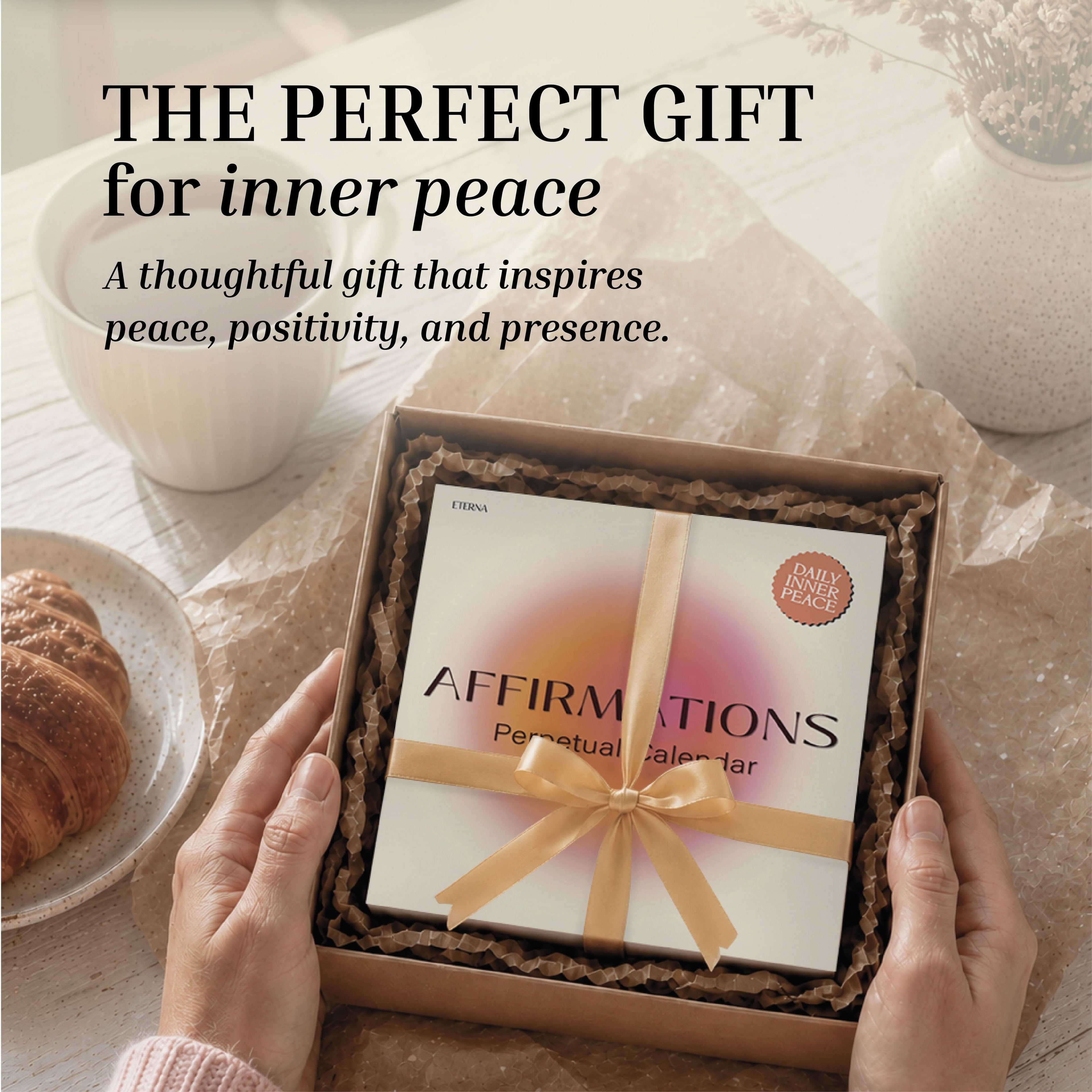 A person holds a gift box featuring the Eterna Goods Perpetual Affirmations Calendar, beautifully tied with a ribbon to offer daily inspiration, while a croissant and a vase sit on the table nearby.