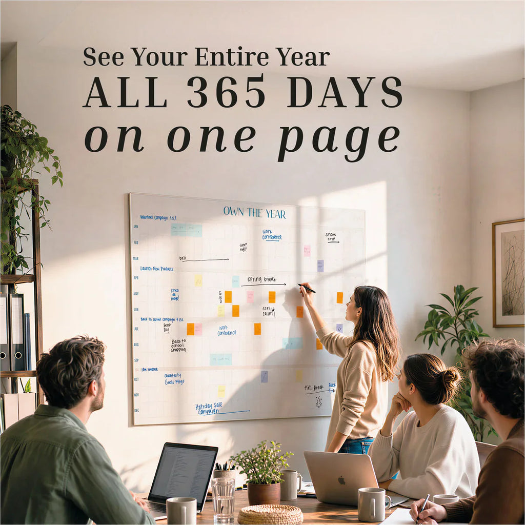 Four people sit at a table with laptops and papers, looking at a large Own The Year Calendar dry erase wall calendar by Eterna Goods as one person writes on it. Text above reads, “All 365 days on one page.”.