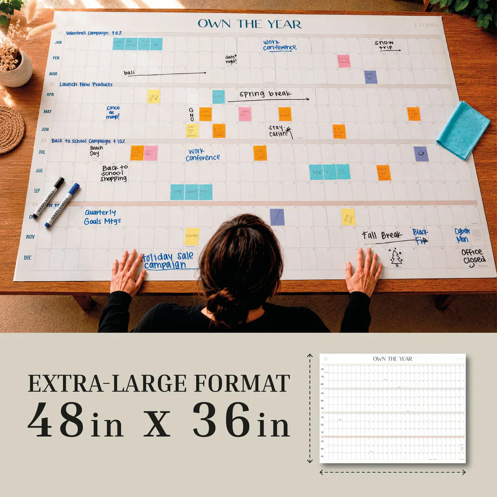 A person sits at a table with the Own The Year Calendar by Eterna Goods, a giant 48x36-inch dry erase wall calendar. Dry erase markers and sticky notes are neatly arranged on the undated calendar, making it an ideal academic wall planner for organizing schedules throughout the year.