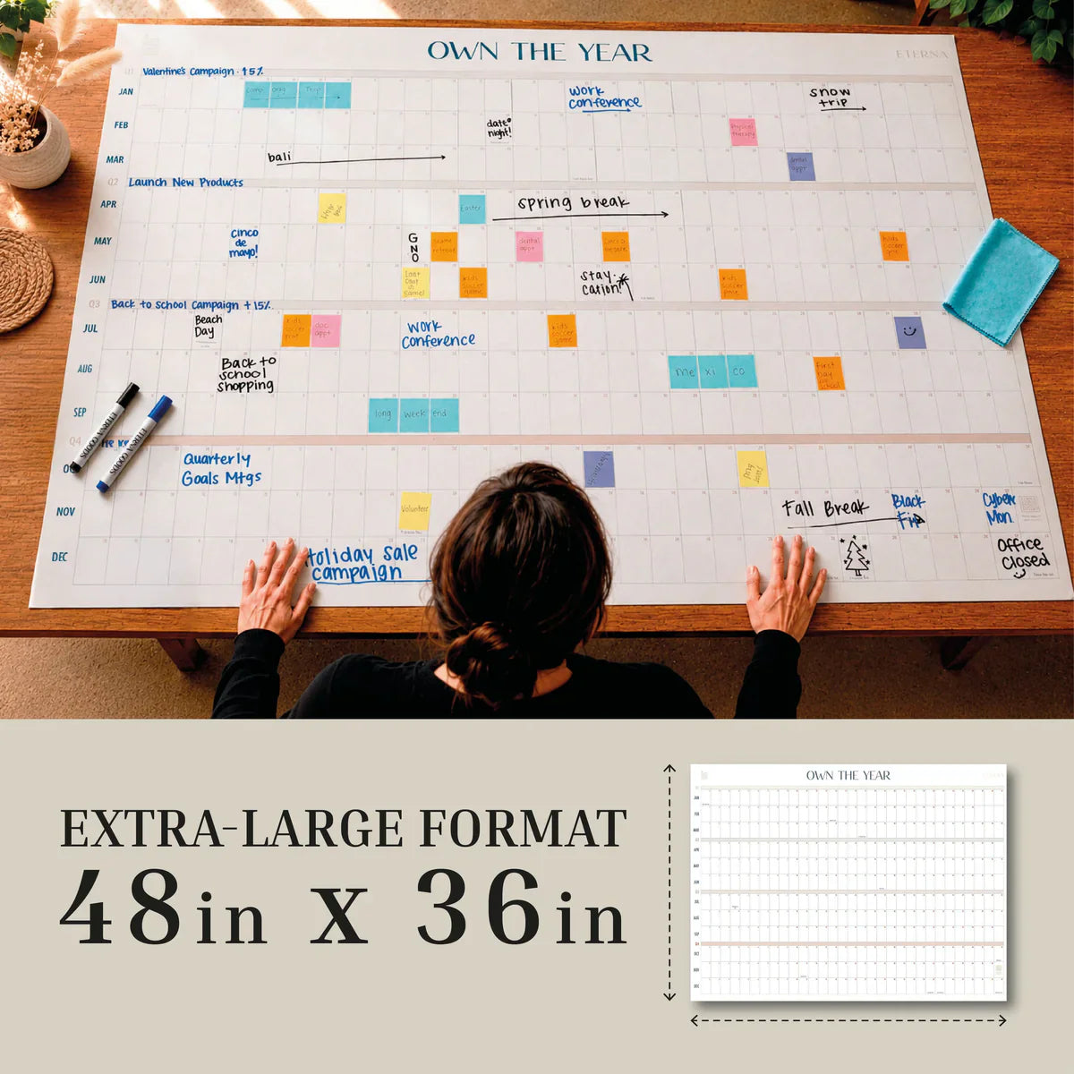 A person sits at a table with the Own The Year Calendar by Eterna Goods, a giant 48x36-inch dry erase wall calendar. Dry erase markers and sticky notes are neatly arranged on the undated calendar, making it an ideal academic wall planner for organizing schedules throughout the year.
