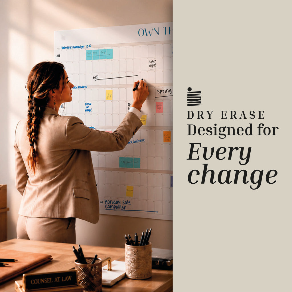 A woman in business attire writes on an Undated Own The Year Calendar by Eterna Goods; text reads “Dry Erase Designed for Every Change.”.