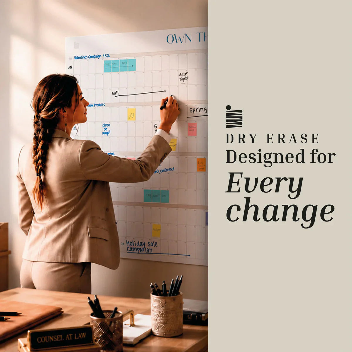 A woman in business attire writes on an Eterna Goods Own The Year Calendar, a wall-mounted giant dry erase wall calendar in an office, with the words “Dry Erase Designed for Every Change” displayed beside her.