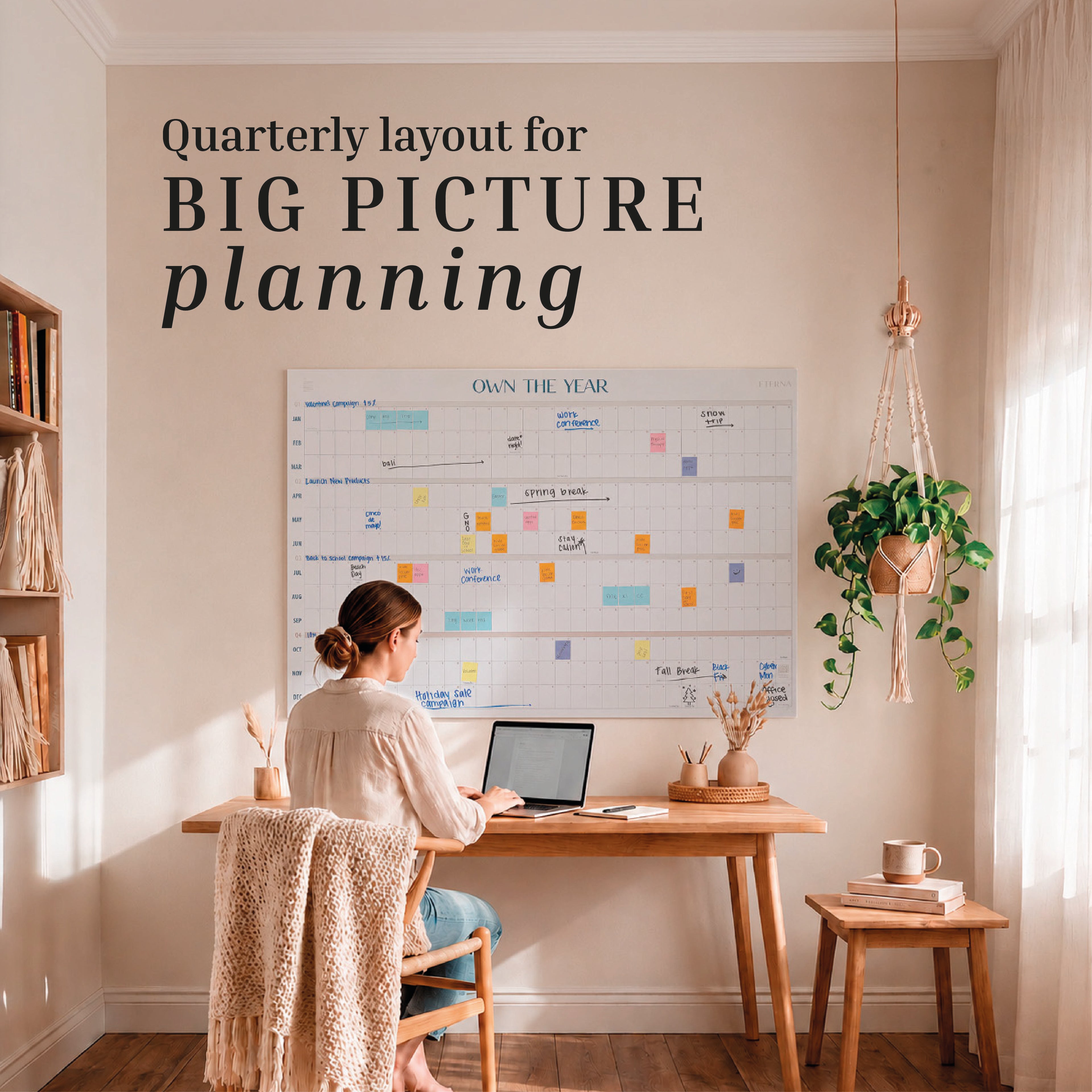 A woman works at a laptop in her cozy, sunlit home office, facing the Eterna Goods Undated Own The Year Calendar—a large reusable dry erase planner—mounted on the wall above her wooden desk.