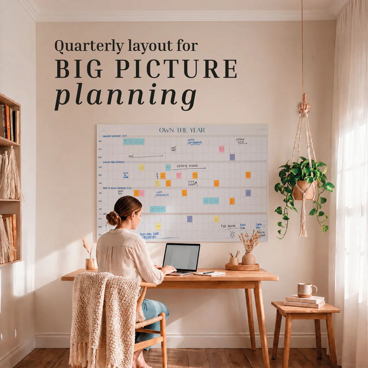 A woman works at her desk on a laptop in a home office, facing an Own The Year Calendar by Eterna Goods—a giant dry erase wall calendar with the words “Quarterly layout for BIG PICTURE planning” displayed prominently.