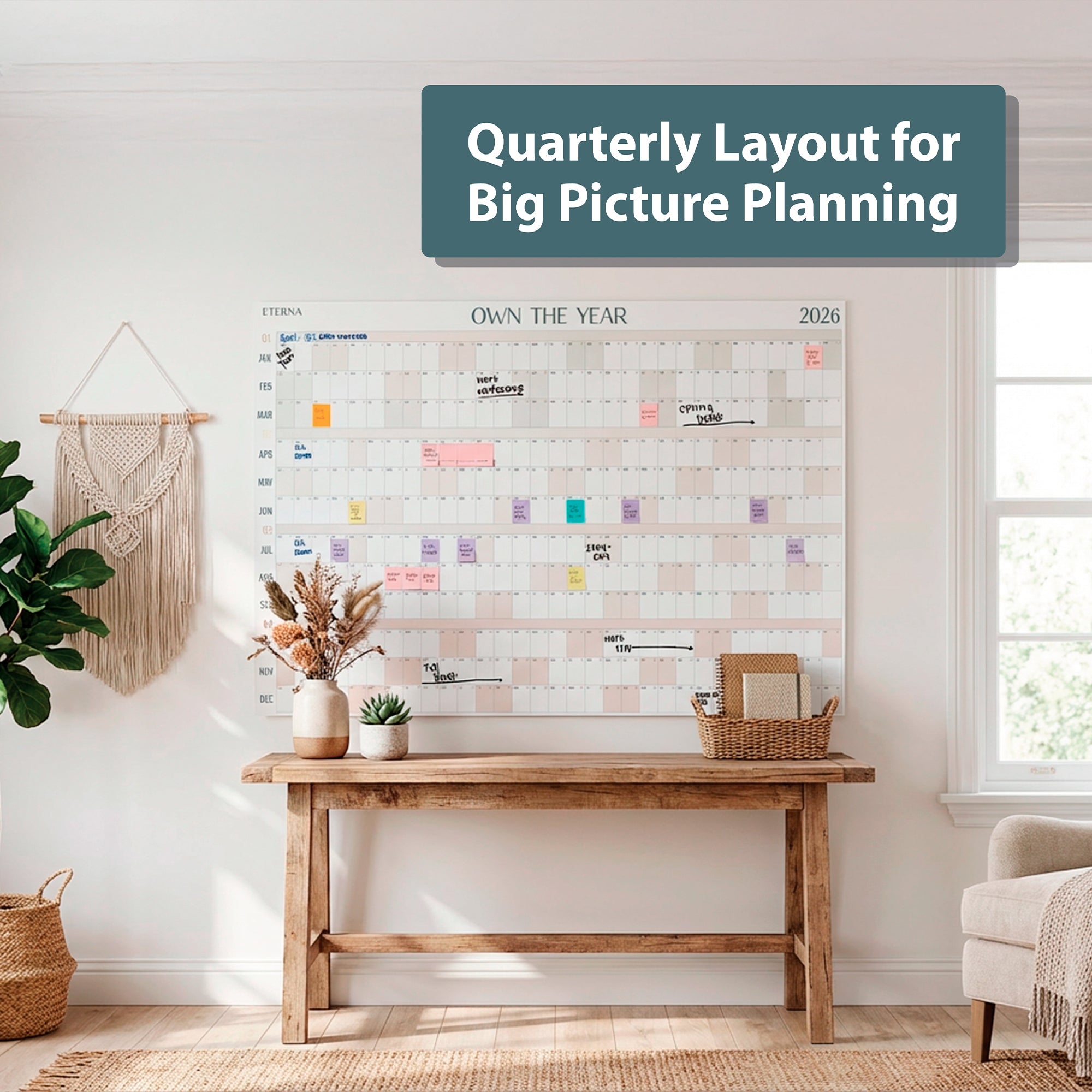 A wooden table with decor sits beneath the Eterna Goods Own the Year 2026 GIANT Dry Erase Wall Calendar (4ft x 3ft, quarterly layout) in a bright, minimal room. A sign reads, "Quarterly Layout for Big Picture Planning.