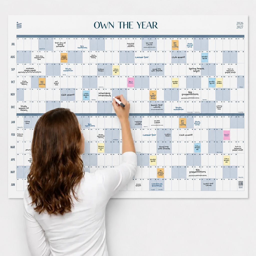 A woman writes on the Own The Year Midyear / Academic Calendar by Eterna Goods—a giant dry erase wall calendar, color-coded and packed with events and reminders, covering July through June.