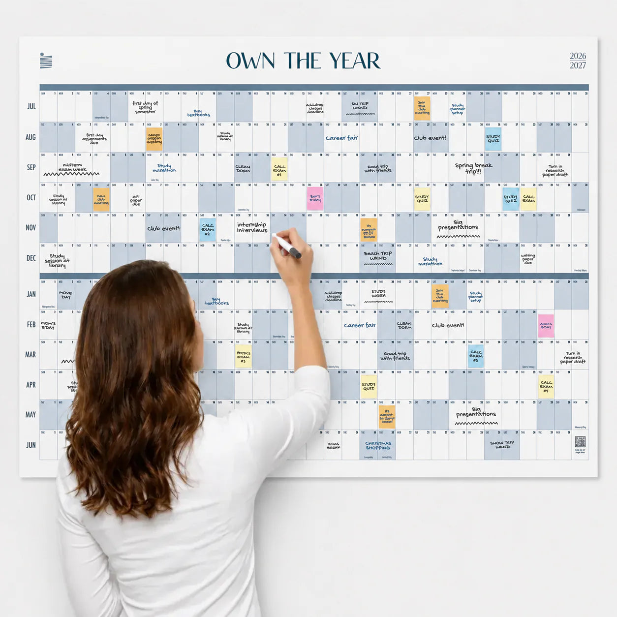 A woman writes on the Own The Year Giant Dry Erase Calendar by Eterna Goods, a large-format wall calendar for 2023-2024 that’s covered in vibrant notes and highlights. This full-year planner helps her stay organized and inspired throughout the year.