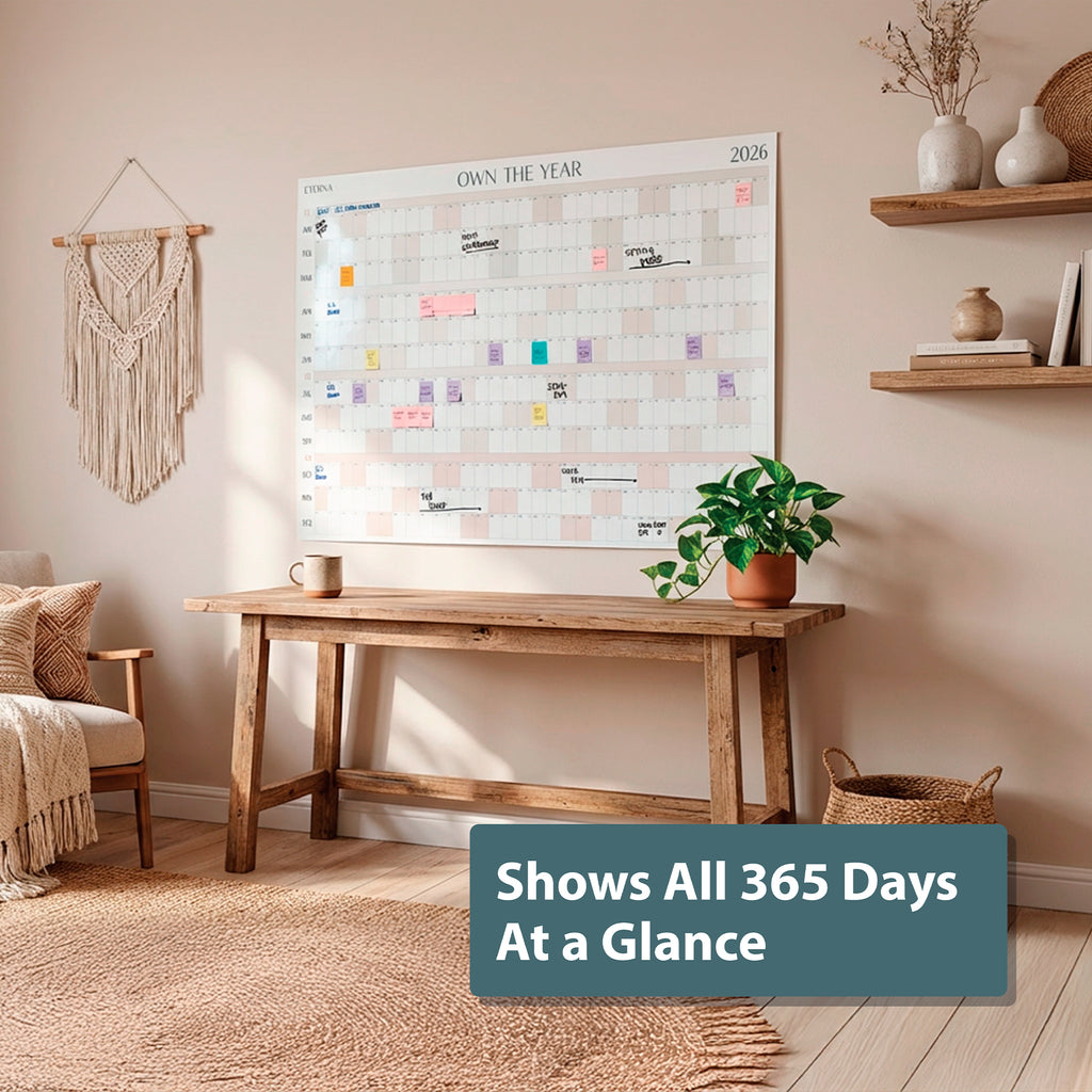 A wooden desk sits against a beige wall, above which hangs the Eterna Goods "Own the Year 2026" GIANT Dry Erase Wall Calendar (4ft x 3ft); plants and woven accents decorate the space. Text overlay: “Shows All 365 Days At a Glance.”.