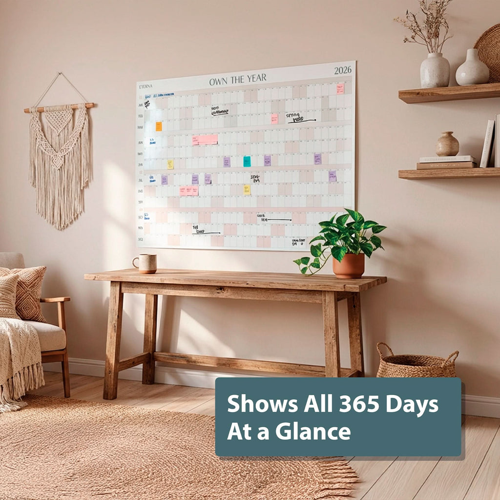 An Own The Year Calendar by Eterna Goods—a giant dry erase wall calendar displaying every day of 2026—hangs above a wooden desk with a plant and mug; a label reads, “Shows All 365 Days At a Glance.”.