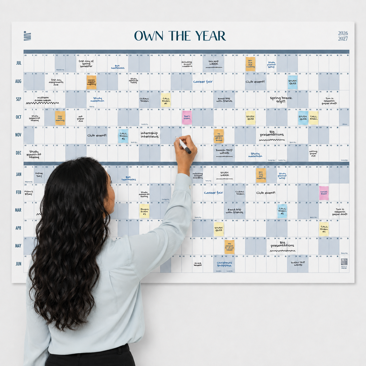 A woman writes on the Eterna Goods 2026-2027 Midyear/Academic Own The Year Calendar, a giant wall calendar covered in multicolored notes and events spanning July 2026 to June 2027.