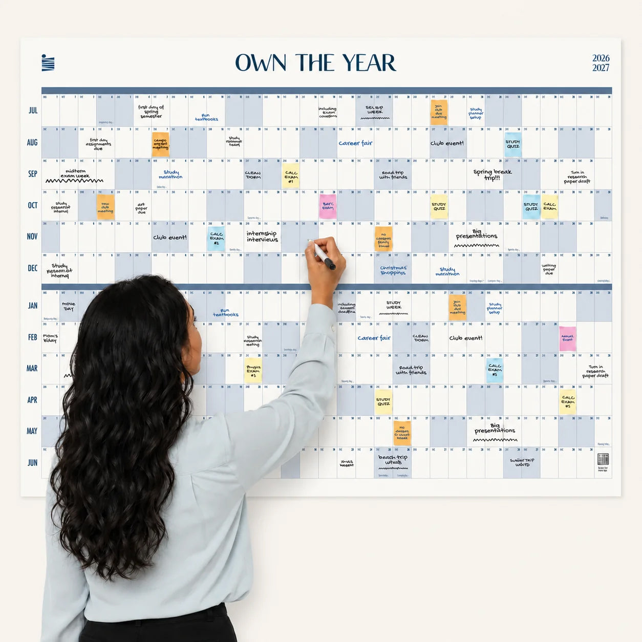 A woman with long hair writes on a large Eterna Goods 2026-2027 Midyear/Academic Own The Year Calendar, which is organized by month and filled with various colored notes and events.