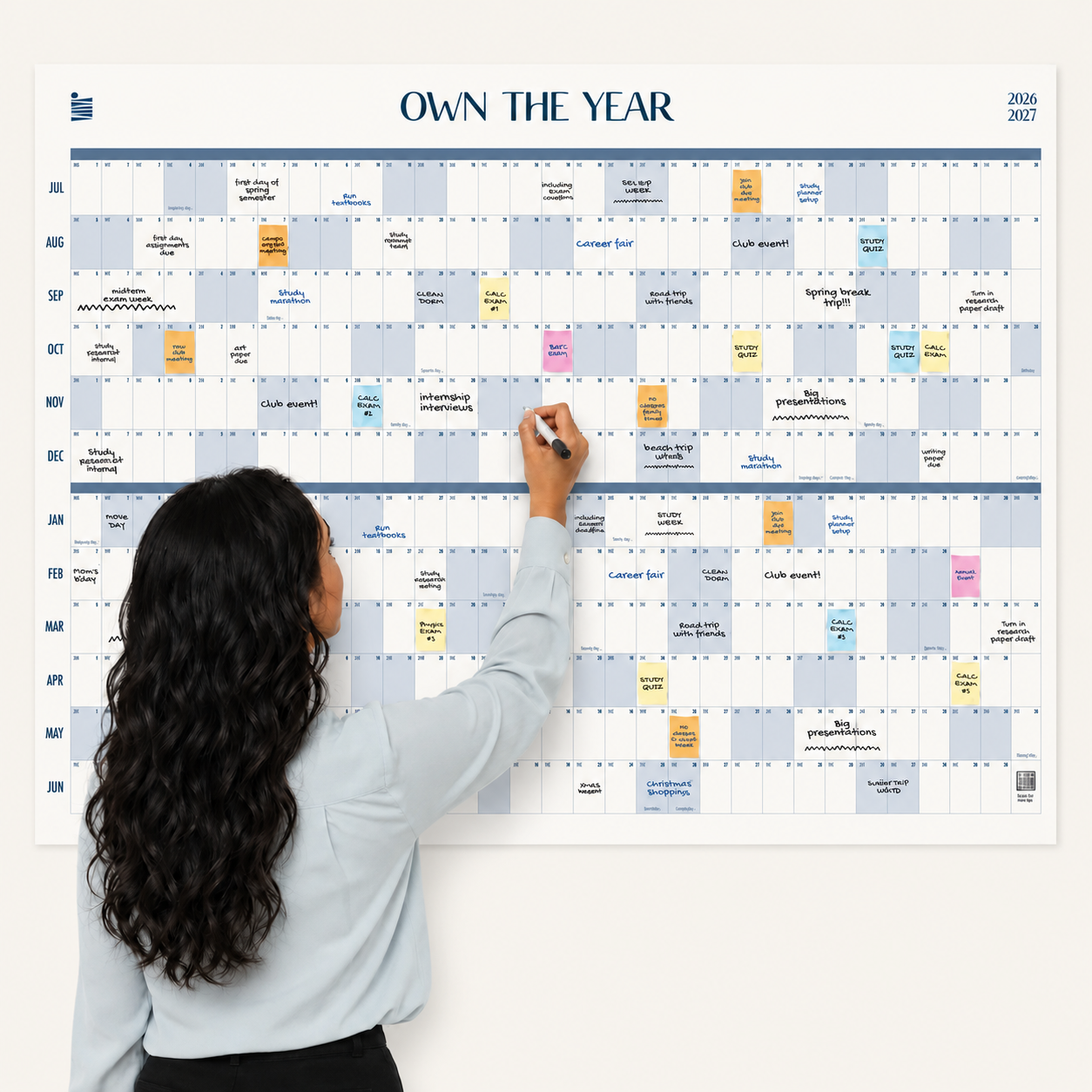 A woman writes on the Eterna Goods 2026-2027 Midyear/Academic Own The Year Calendar, a large dry erase wall calendar covered with colorful sticky notes and events organized from July through June.