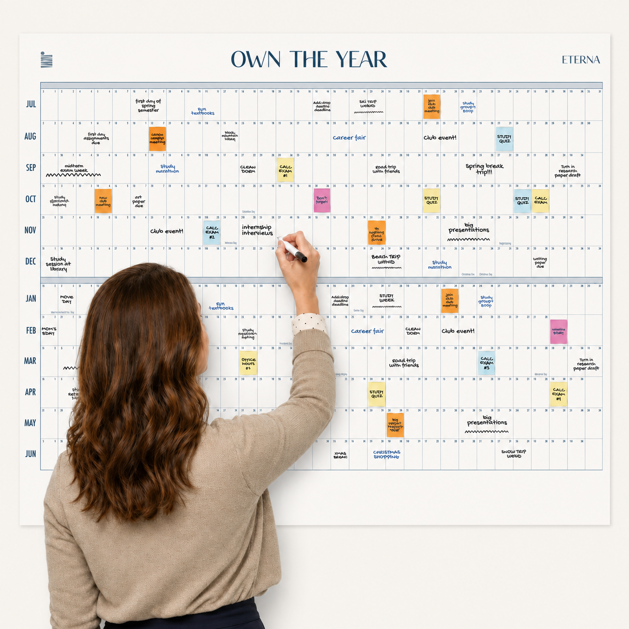 A woman writes on the Eterna Goods Undated Midyear/Academic Own The Year Calendar, a giant dry erase wall calendar filled with colorful sticky notes and handwritten events, covering a full year from July to June.