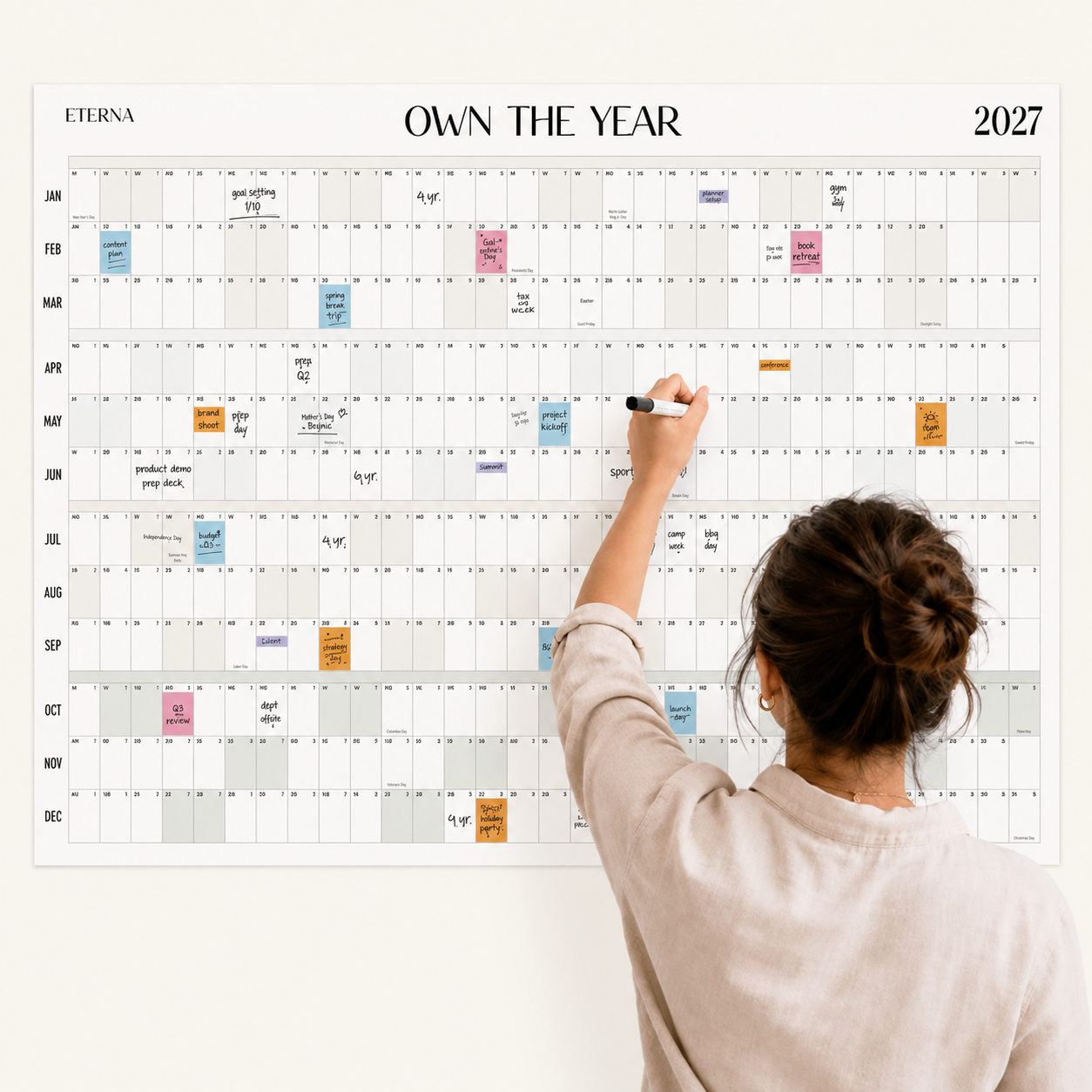 A person writes on the Eterna Goods Dated Own The Year Calendar for 2027, which is covered with colorful sticky notes and planning details.