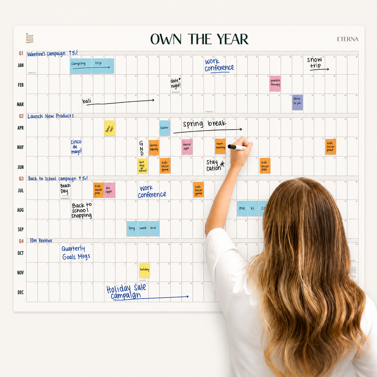 A woman writes on the Undated Own The Year Calendar by Eterna Goods, a wall-mounted yearly planner filled with events, tasks, and notes written in various colors.