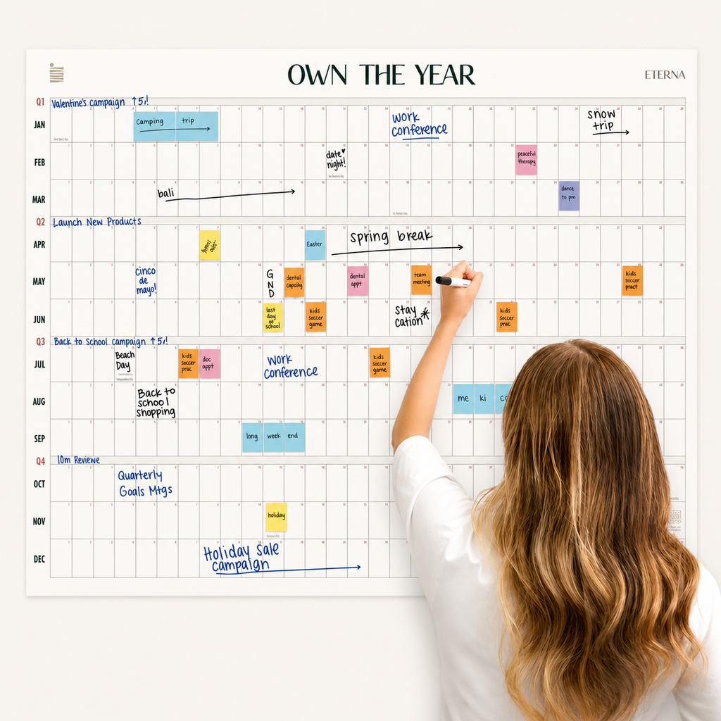 A woman writes on the Undated Own The Year Calendar by Eterna Goods, a wall-mounted yearly planner filled with events, tasks, and notes written in various colors.