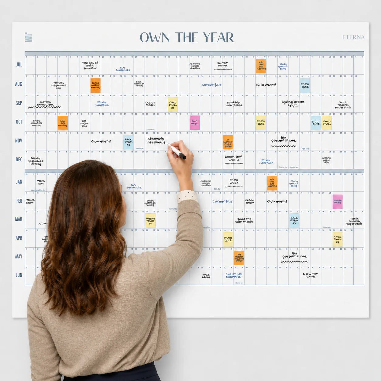 A woman writes on the Eterna Goods Own The Year Midyear / Academic Calendar, a giant wall planner filled with colorful sticky notes and handwritten events that covers a full year from July to June.