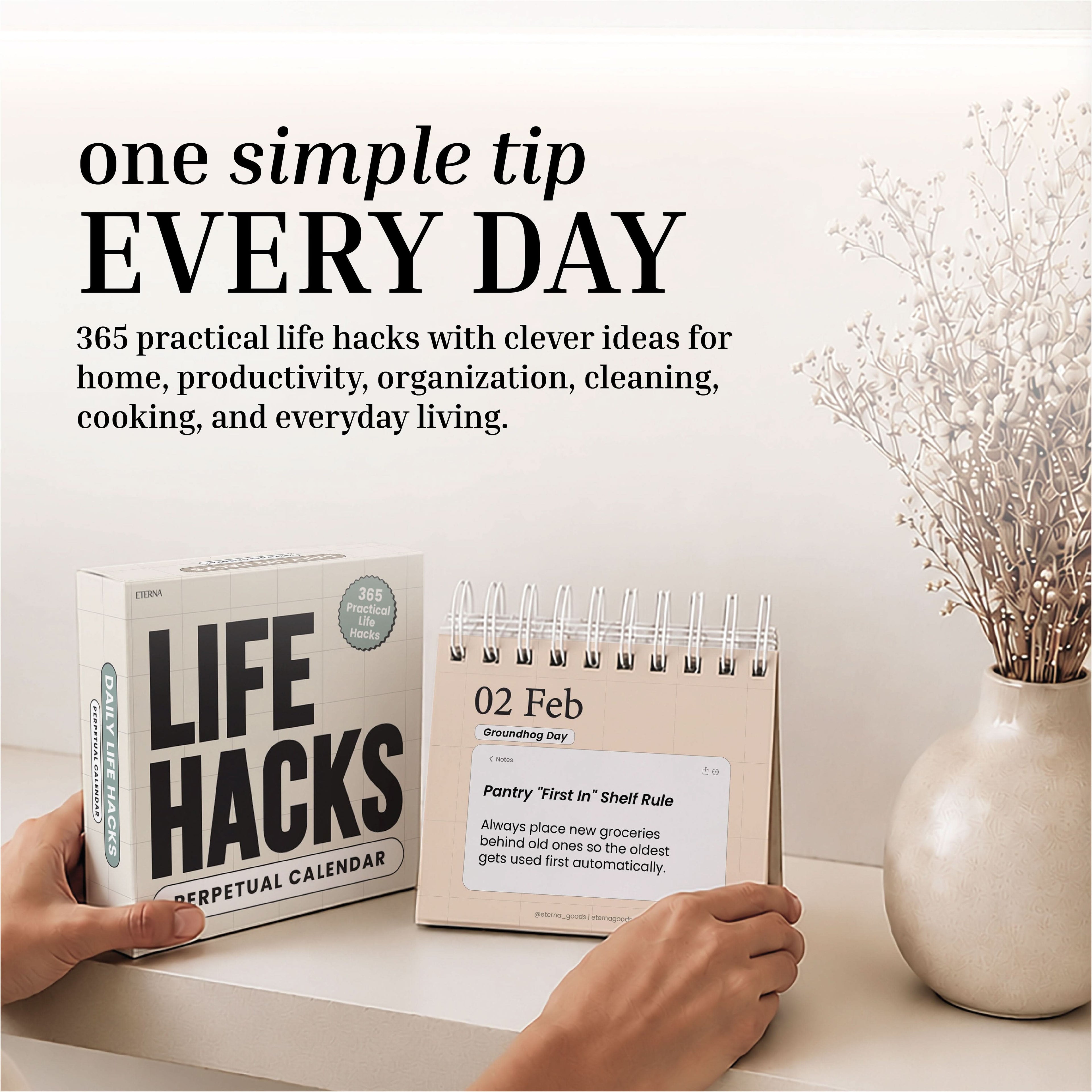 A hand flips a page on the Eterna Goods Perpetual Life Hacks Calendar, a stylish desktop calendar that delivers daily productivity tips. The scene is set against a backdrop of neutral decor and a vase filled with dried flowers.
