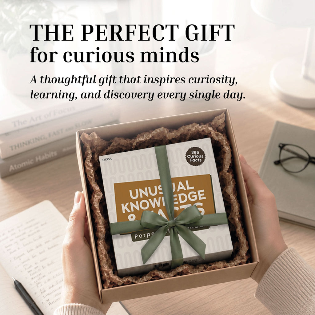 Hands hold an open gift box containing Eterna Goods’ "Perpetual Unusual Knowledge & Facts Perpetual Calendar," accented with a green ribbon—an ideal trivia gift. Books and glasses are on the table in the background.