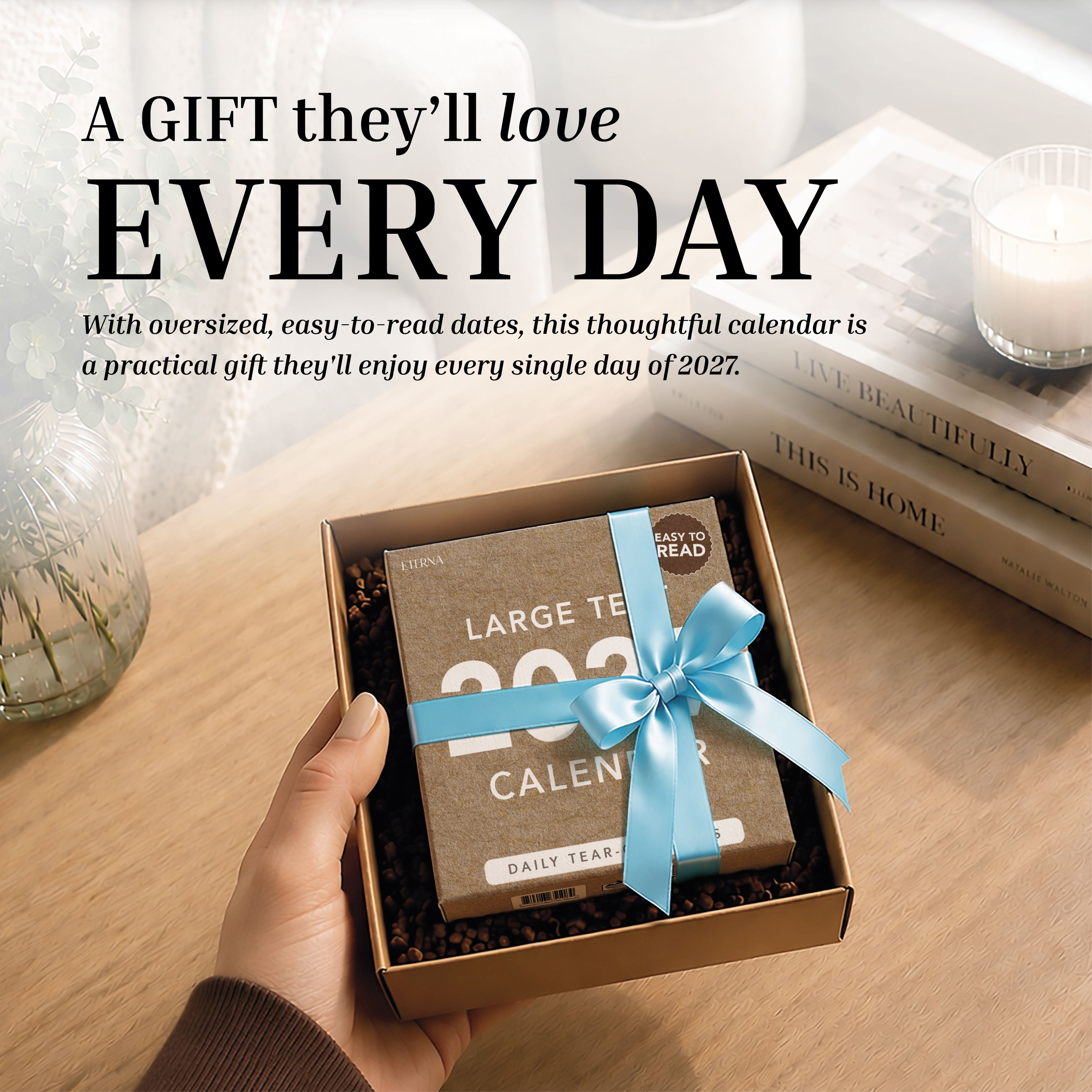 A person holds an open gift box containing an Eterna Goods 2027 Large Text Calendar—a daily tear-off desk calendar with easy-to-read dates—tied with a blue ribbon; a candle and cup rest on the table.