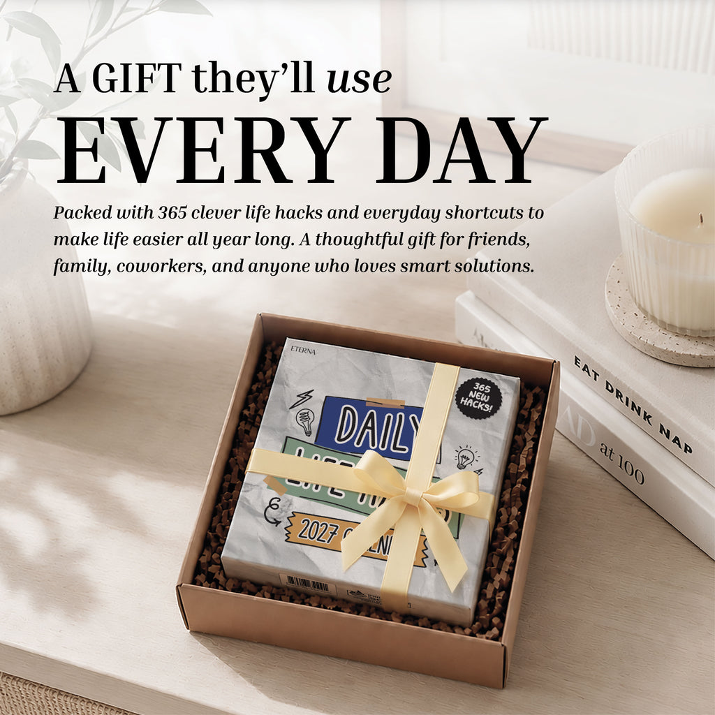 A boxed gift with a ribbon, containing the Eterna Goods "2027 Daily Life Hacks Calendar"—a tear-off desk calendar filled with productivity tips and daily hacks—sits on a table next to a lit candle and a book titled "Eat Drink Nap.