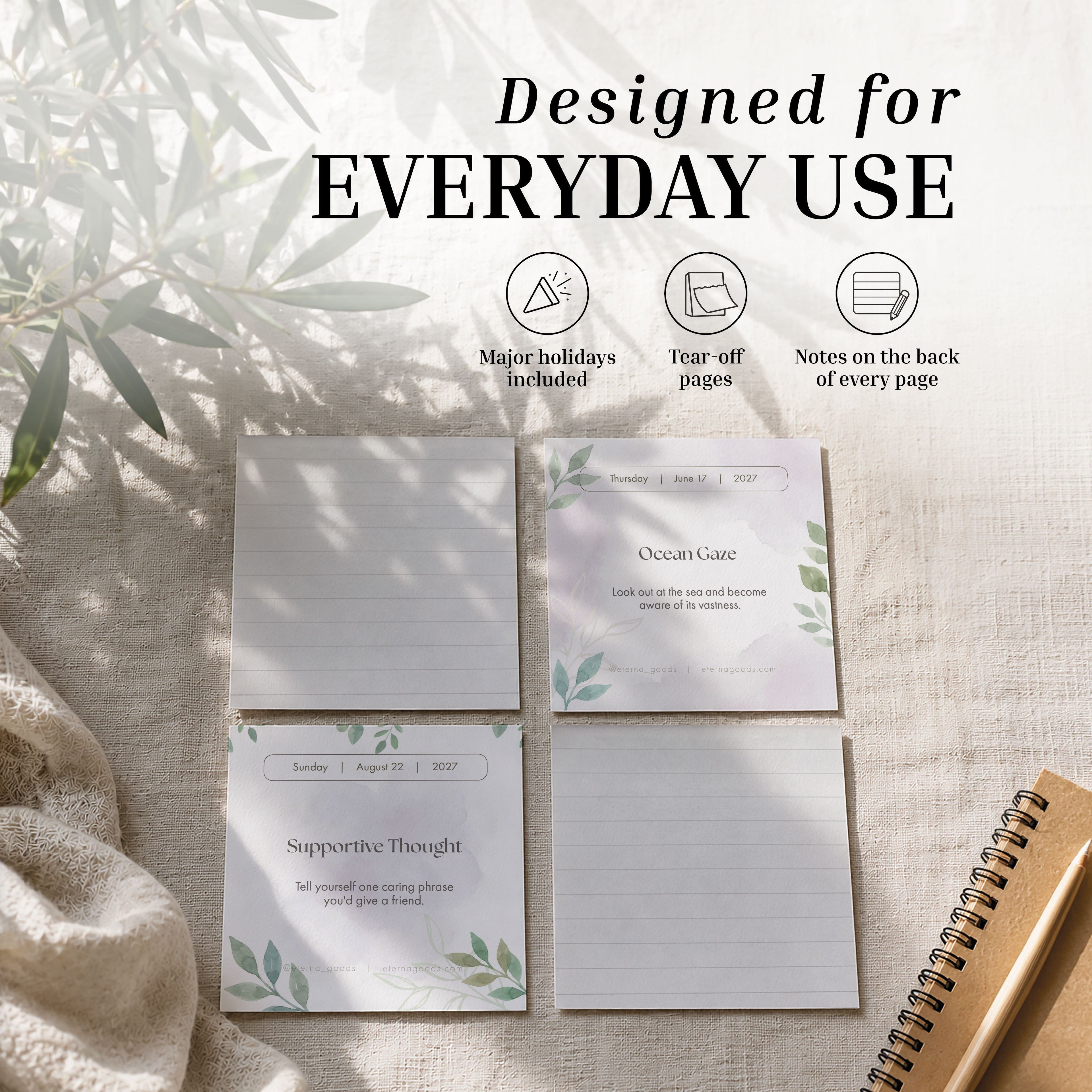 On a beige fabric surface, three pages from the Eterna Goods 2027 Mindfulness Calendar feature dated prompts for daily mindfulness exercises, supportive thoughts, and lined space for notes—displayed next to a spiral notebook and bathed in sunlit shadow.