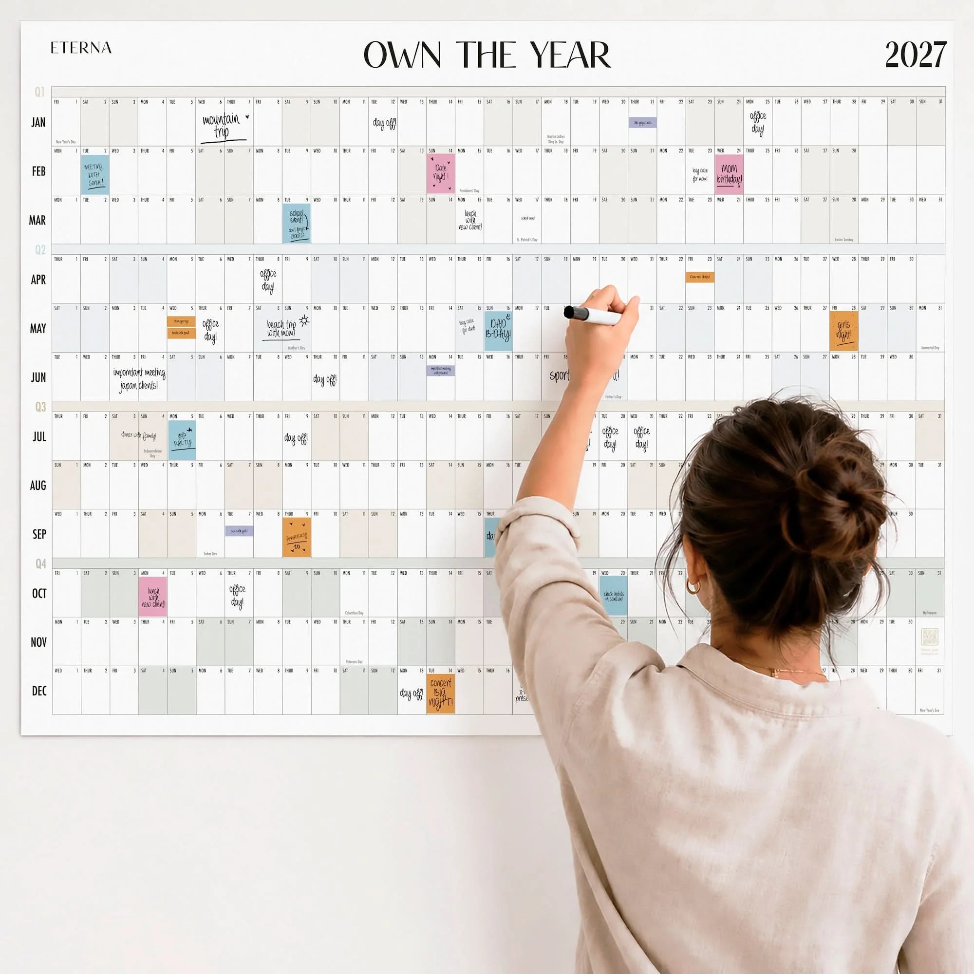A woman writes on the Own The Year Calendar by Eterna Goods—a giant dry erase wall planner for 2027 that spans the entire year and covers the wall, with dates and events highlighted in different colored boxes.