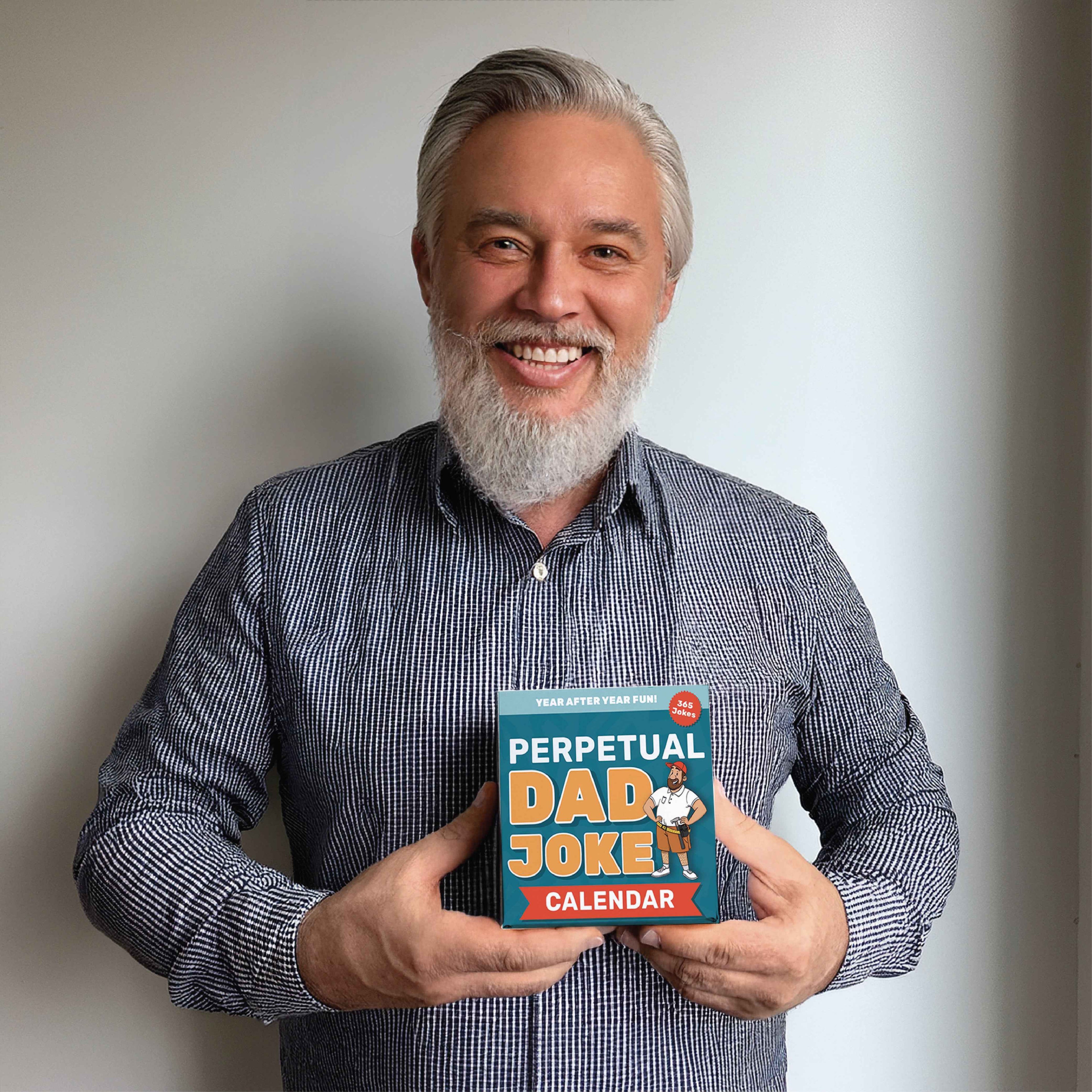 A smiling man with gray hair and beard holds the Eterna Goods Perpetual Dad Joke Calendar, featuring full-color art, against a plain white background.