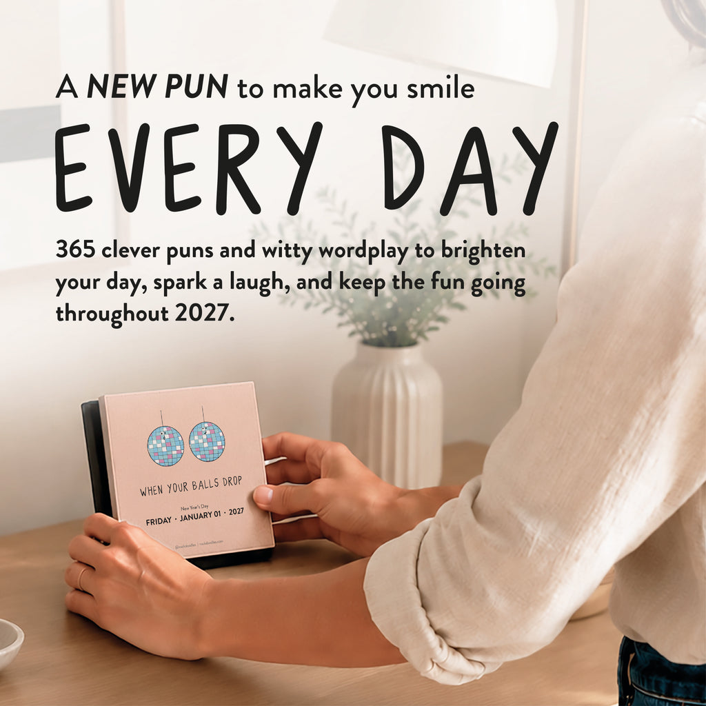A person flips through the 2027 A Pun a Day Calendar by rockdoodles on their desk beside a vase and lamp, with bold text highlighting clever wordplay for every day of the year.