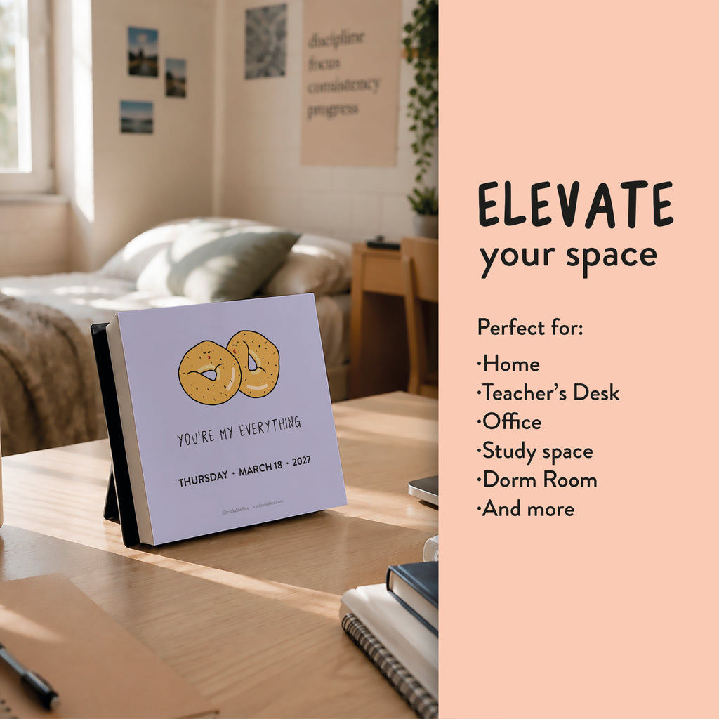 The 2027 A Pun a Day Calendar by rockdoodles, featuring a charming bagel illustration and the phrase "YOU'RE MY EVERYTHING," sits on a wooden desk in a sunlit, organized room adorned with plants and wall art in the background.