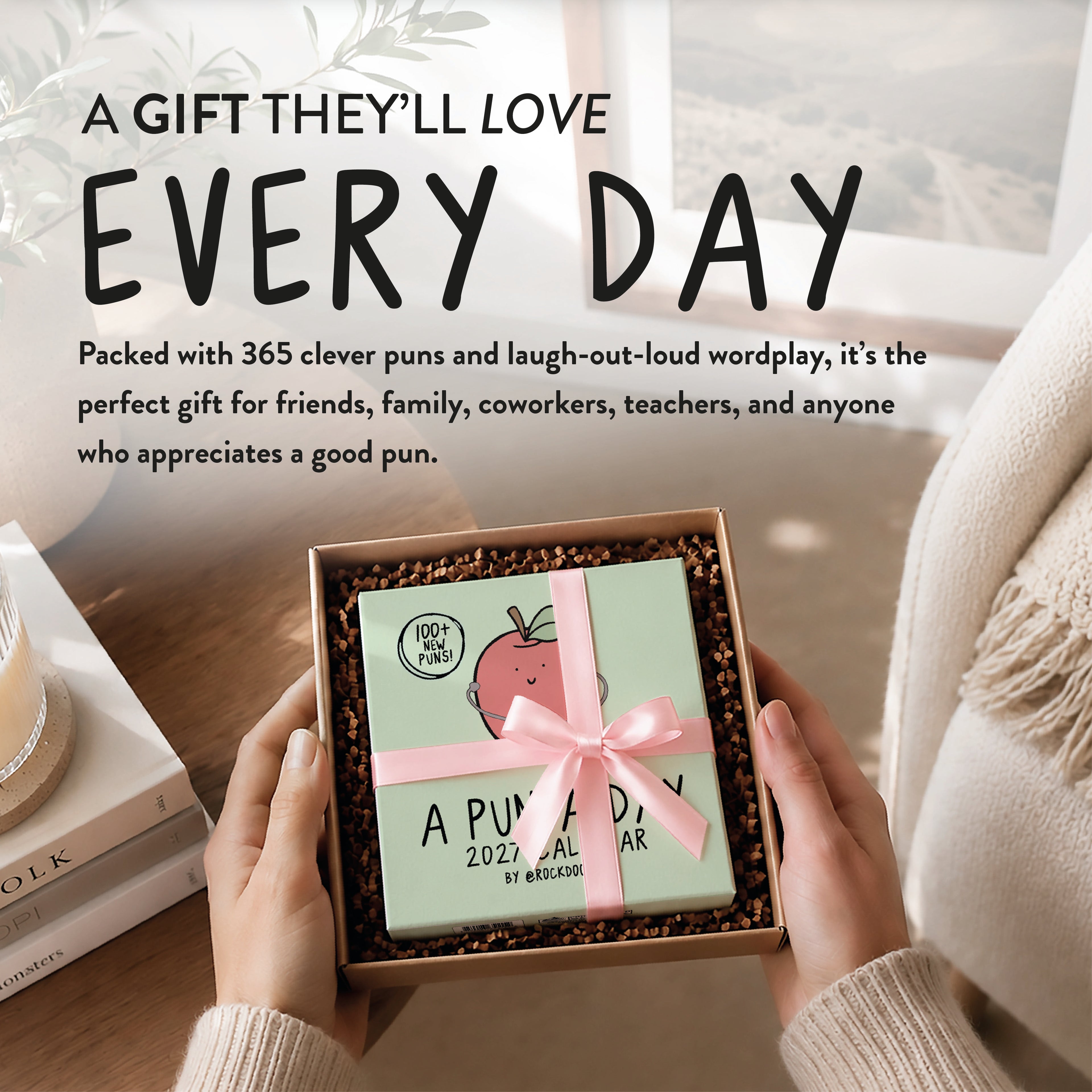 Hands hold an open gift box containing a "2027 A Pun a Day Calendar" by rockdoodles. The box, filled with brown packing material and tied with a pink ribbon, promises endless wordplay and laughter. A cozy, sunlit room is in the background.