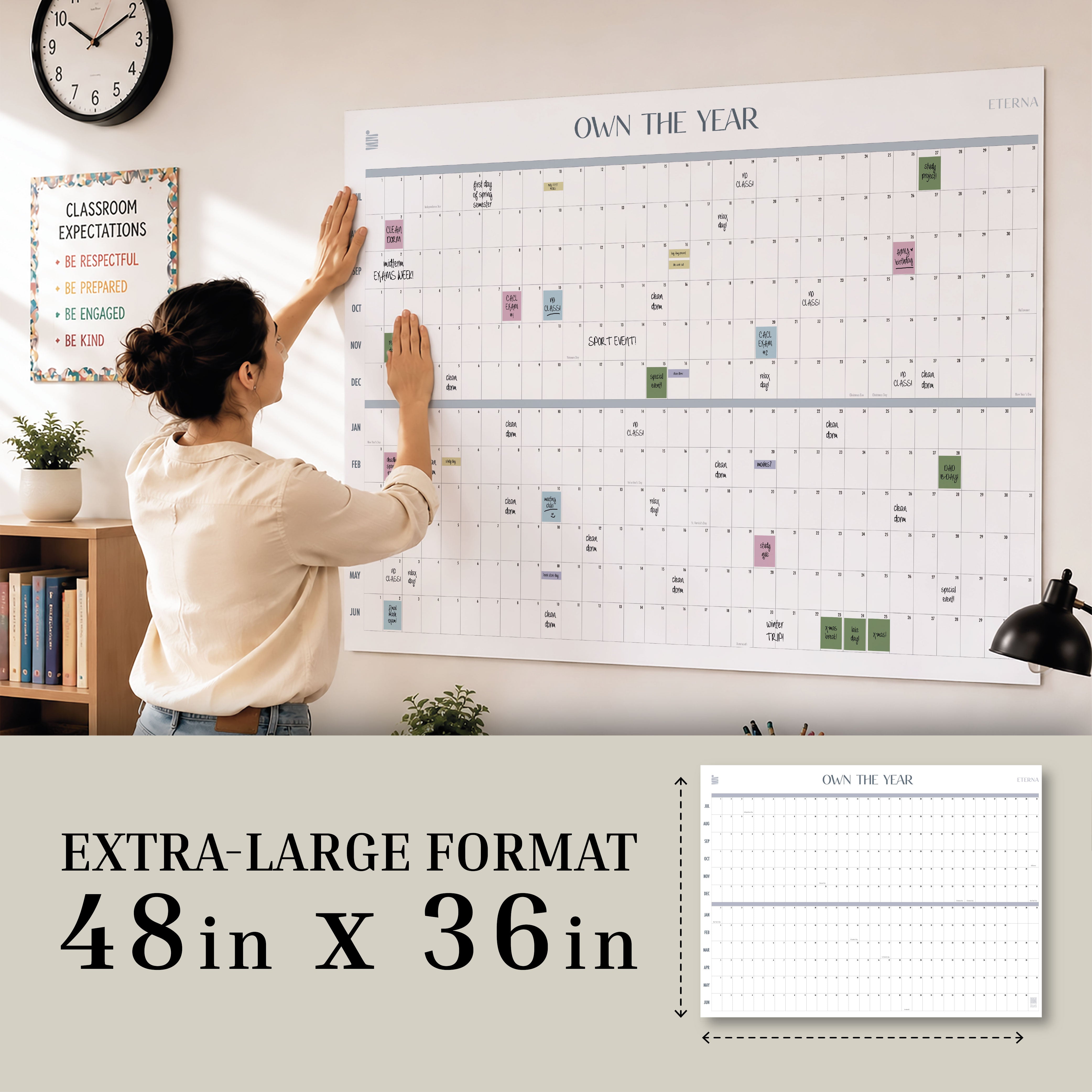 A woman hangs the “Own The Year” Undated Academic Calendar by Eterna Goods, measuring 48x36 inches, on the wall in a home or office; in the background, a classroom expectations sign and another undated academic wall calendar are visible.