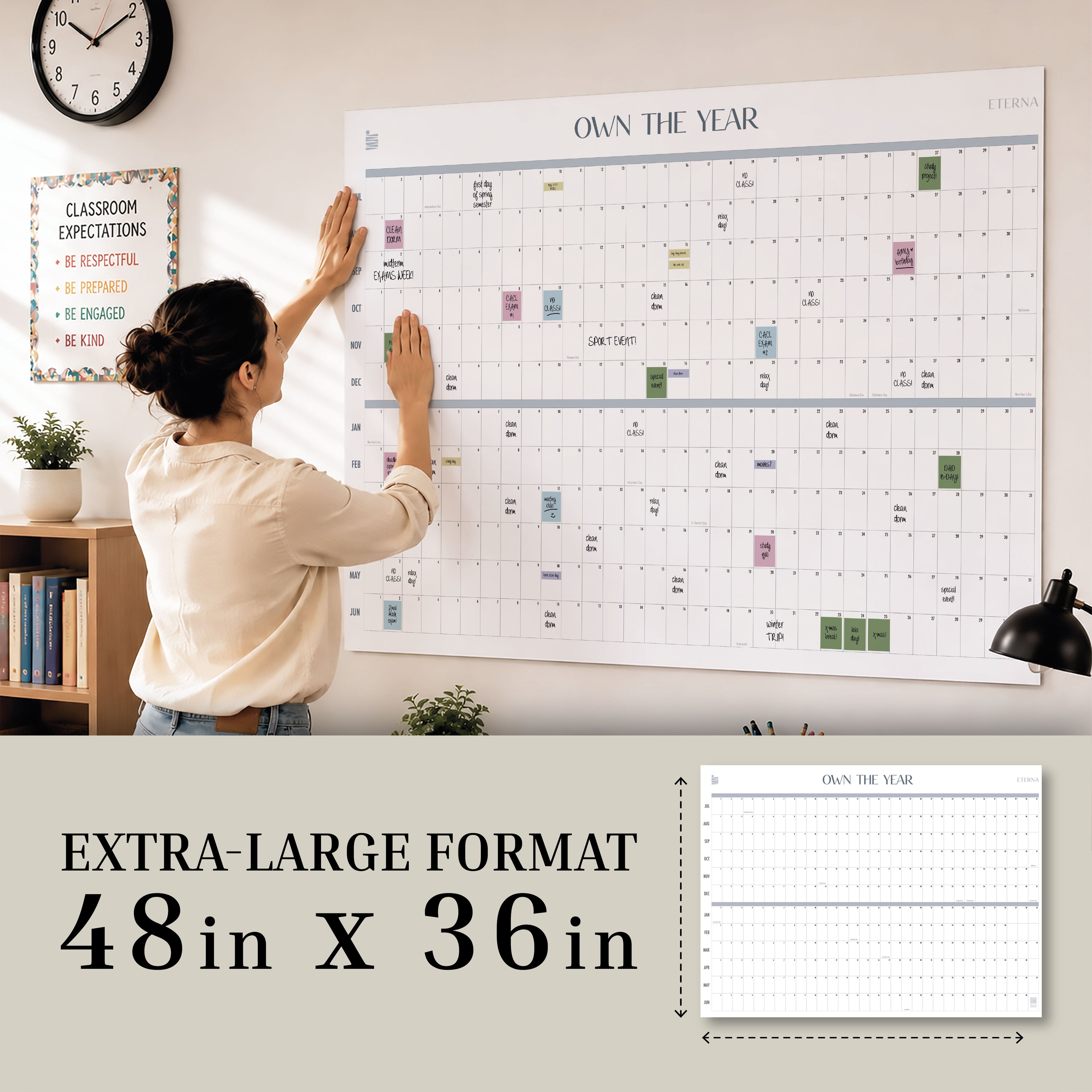 A woman hangs the “Own The Year” Undated Academic Calendar by Eterna Goods, measuring 48x36 inches, on the wall in a home or office; in the background, a classroom expectations sign and another undated academic wall calendar are visible.