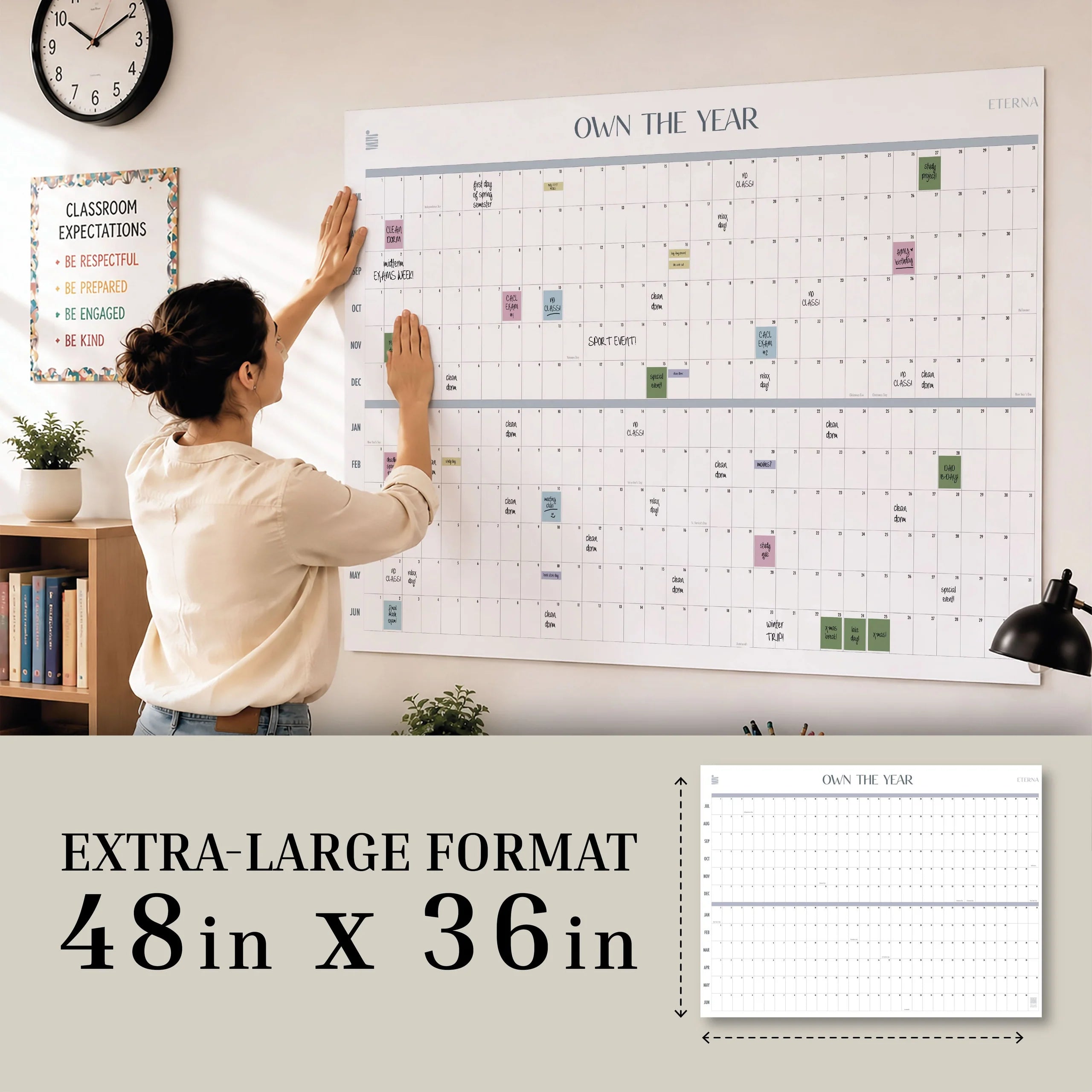 A person mounts an Eterna Goods Own The Year Midyear / Academic Calendar (48x36-inch giant dry erase wall calendar) in a classroom. A plant, lamp, and motivational poster are visible in the background.