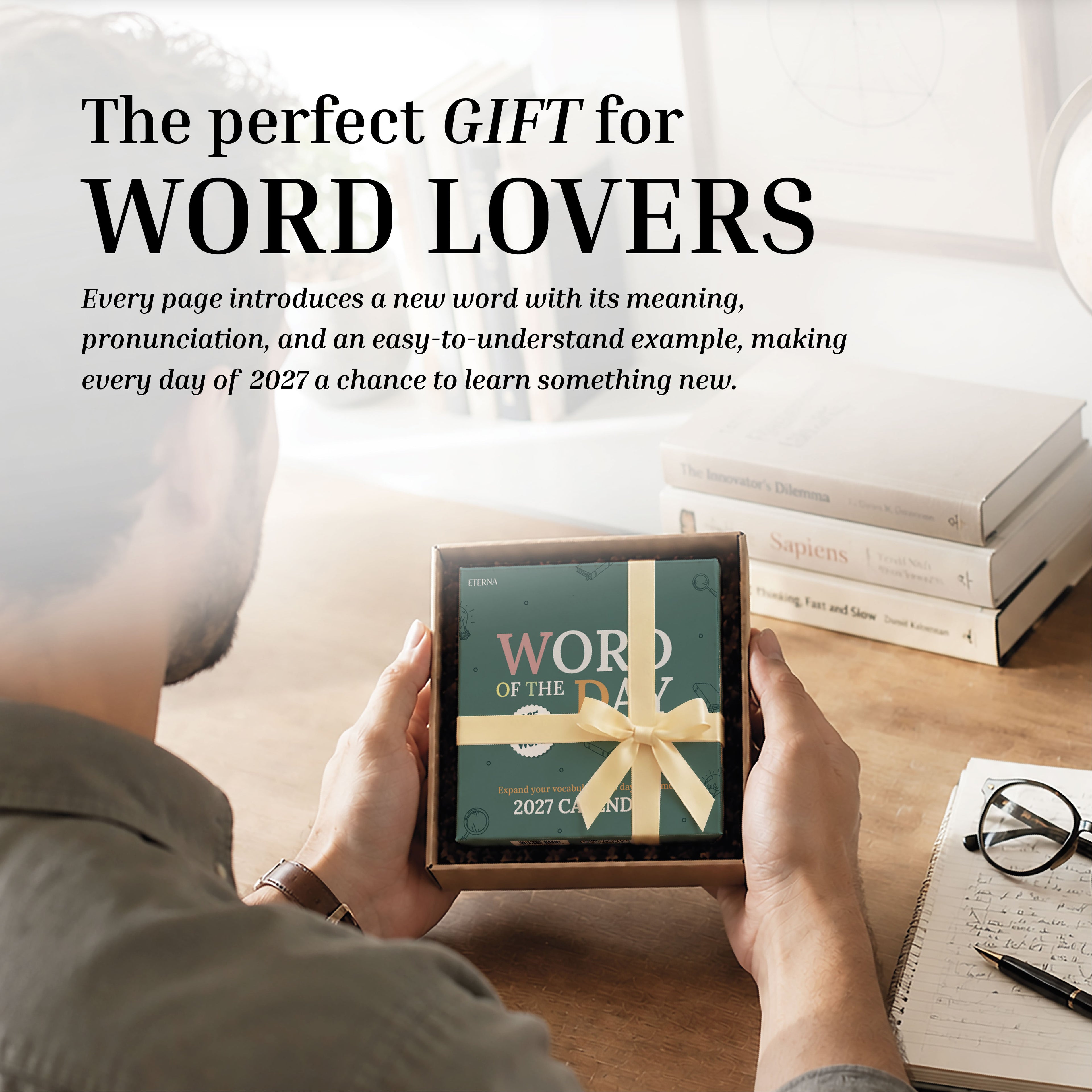 A person sits at a desk with books, glasses, and a coffee cup, holding a ribbon-wrapped Eterna Goods 2027 Word Of The Day Calendar—ready to expand their vocabulary in style.